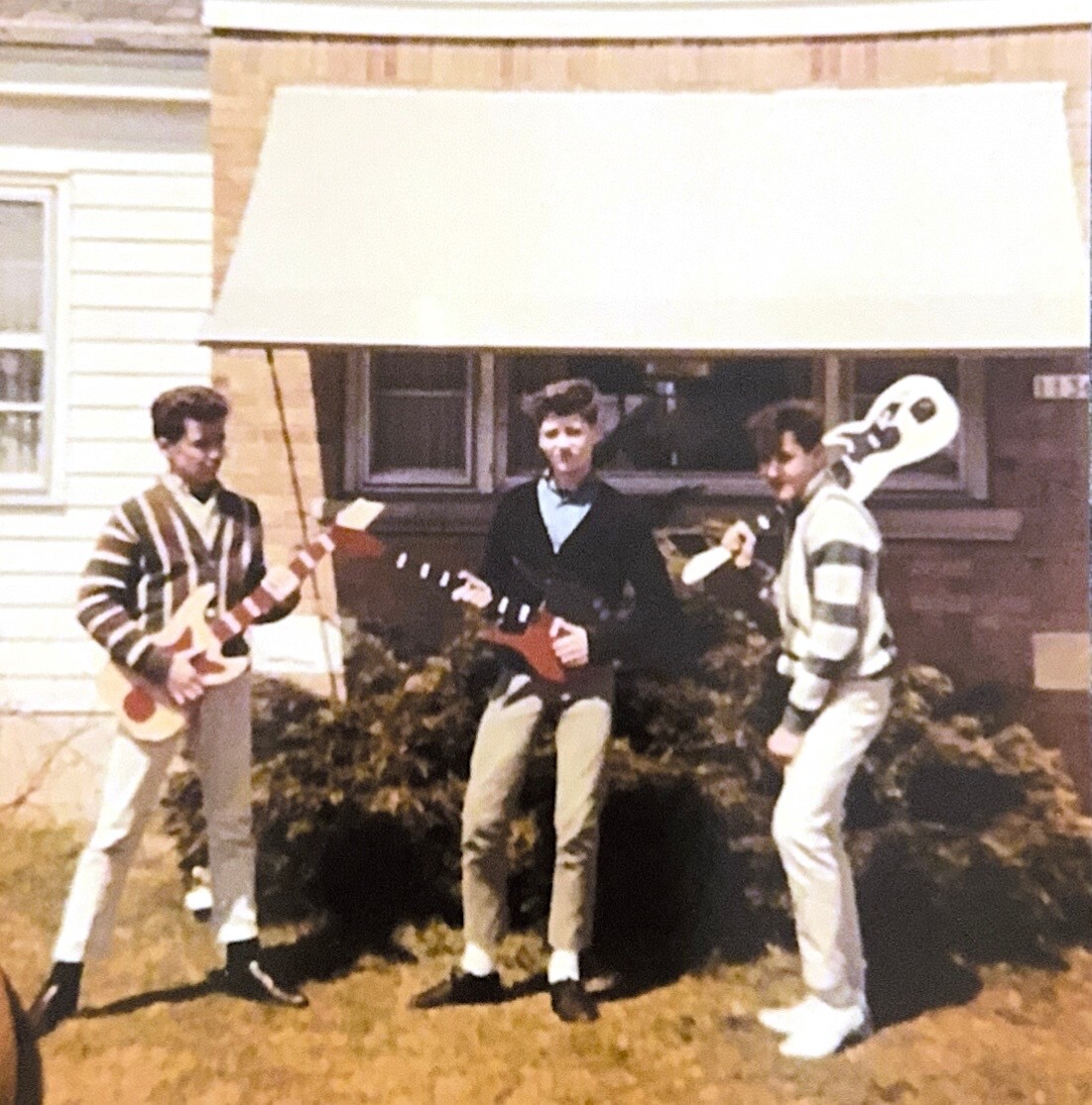 Three teens pose with guitar props outside a house; one wears a white bird mask on their head.