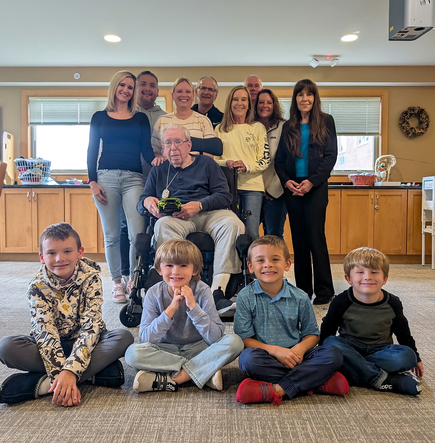Indoor group photo: elderly man in a wheelchair at center, surrounded by smiling adults standing and several children seated on the floor in front.