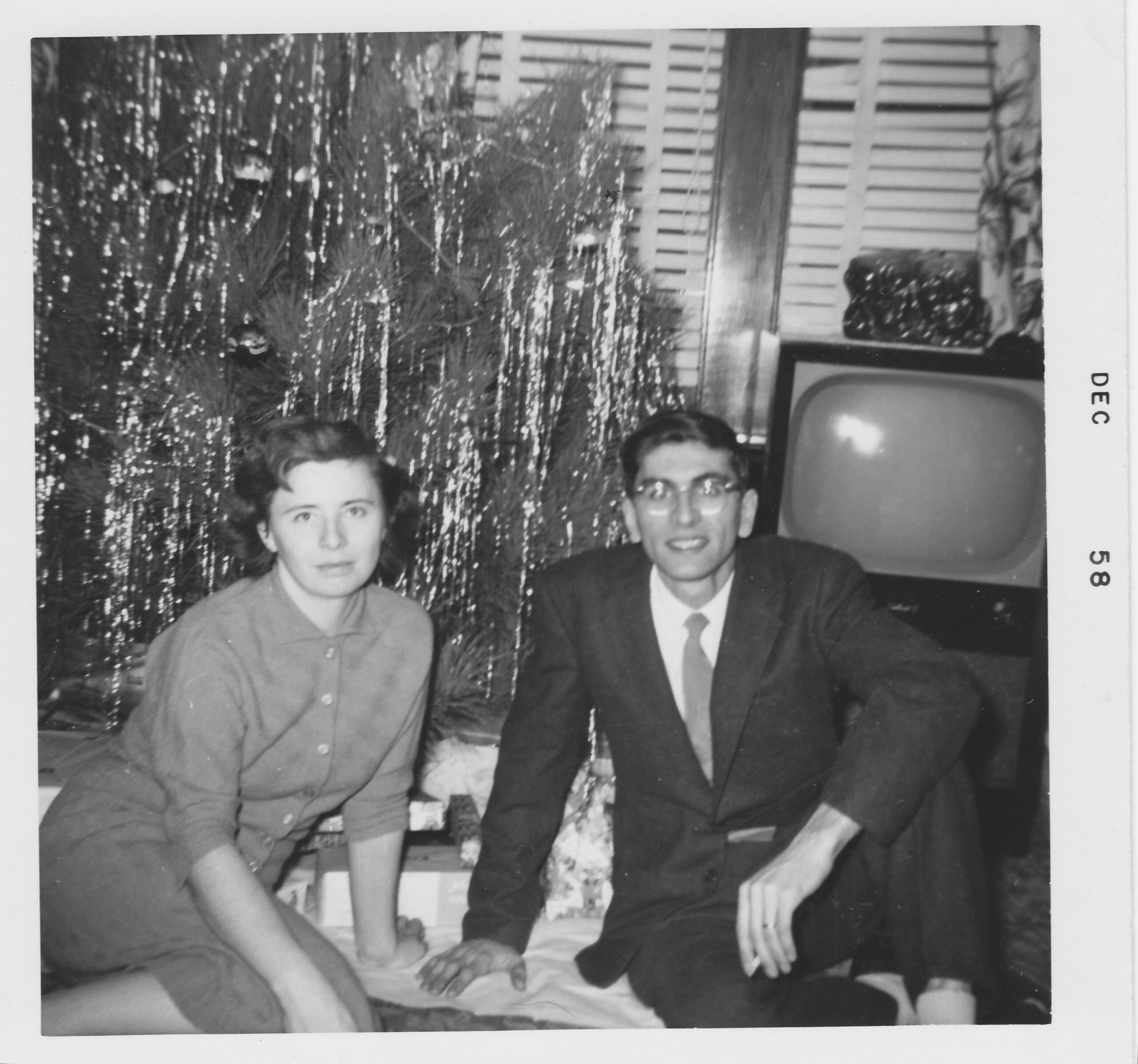 Two people sit together on a couch indoors, a woman on the left and a man in a suit on the right, with a decorated Christmas tree in the background.