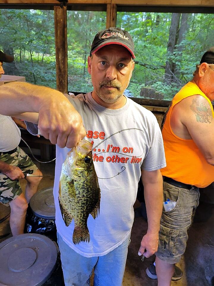 Man in a gray t-shirt and cap holds up a largemouth bass by its mouth inside a covered outdoor shelter, with others in the background.