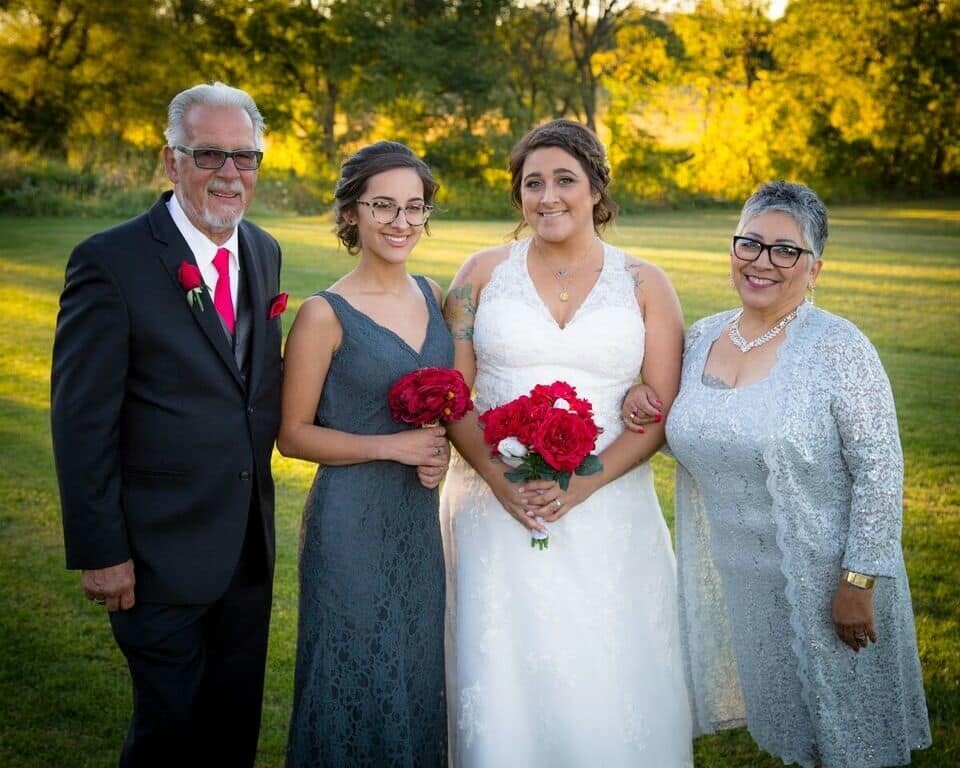 Bride in a white dress holds a red bouquet, flanked by two women in gray dresses and a man in a suit outdoors on a sunny day.