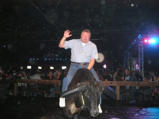Man rides a bucking mechanical bull at a crowded indoor event with stage lighting visible in the background.