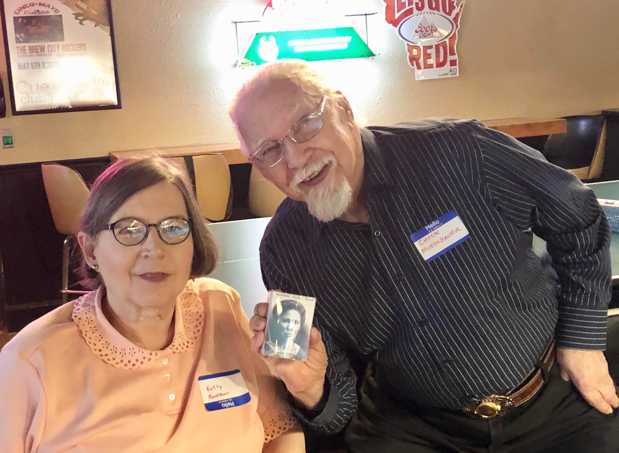 Two smiling seniors pose together in a casual restaurant; the man holds a small black-and-white photo of a man.
