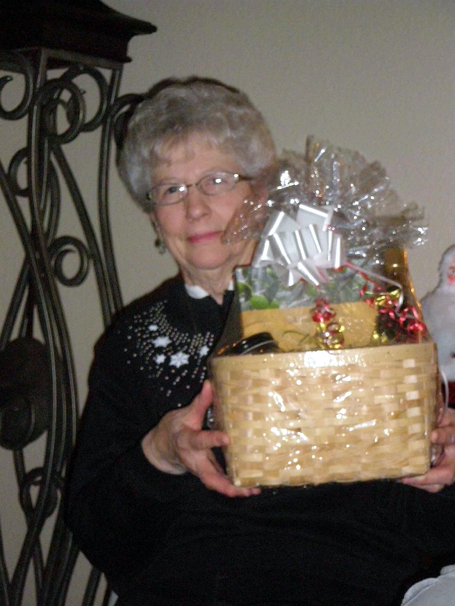 Smiling elderly woman with glasses holds a large wicker gift basket wrapped in clear cellophane and ribbon indoors.
