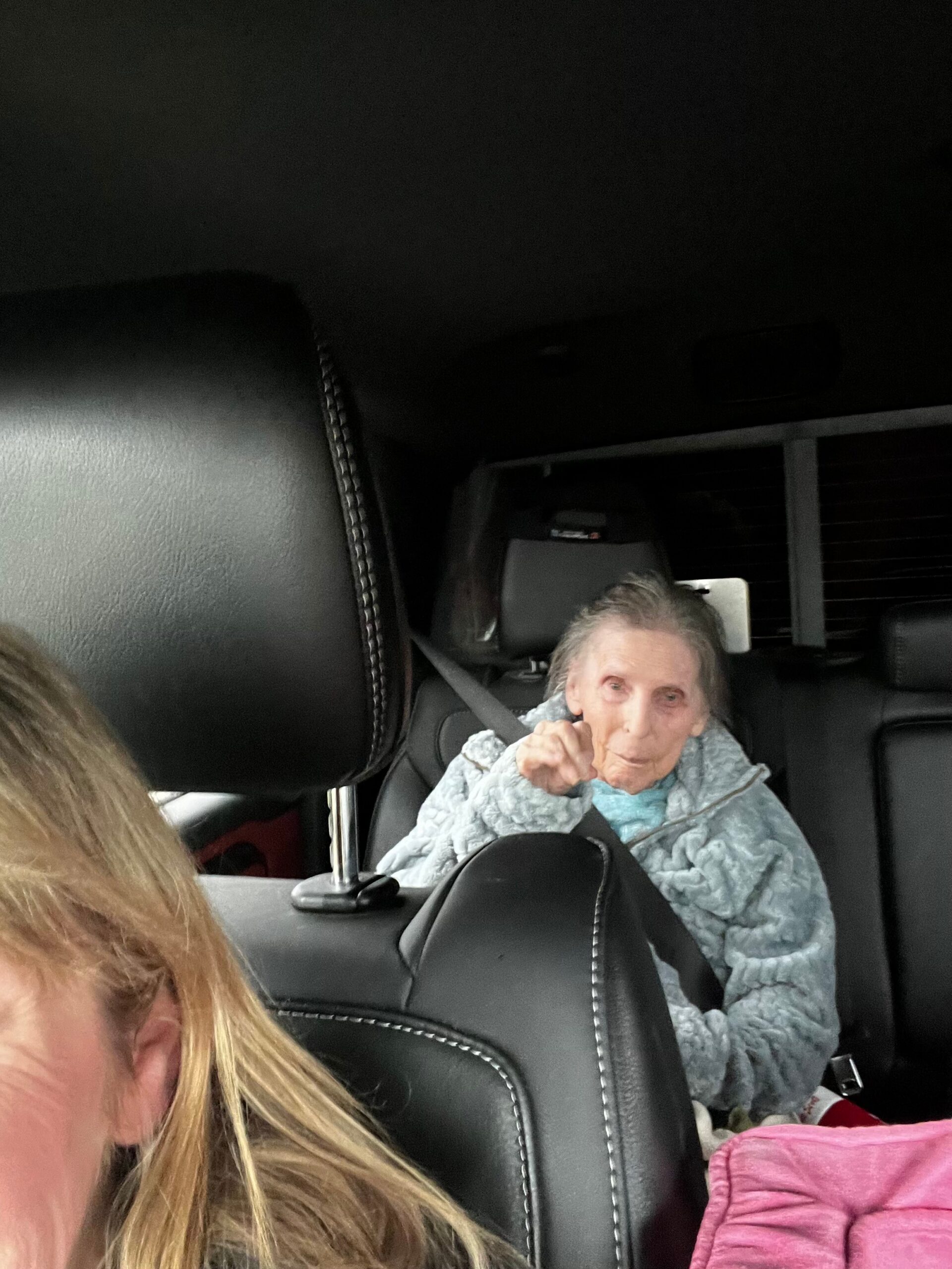 Older woman in the back seat of a car, wrapped in a light blue quilt and wearing a seat belt.