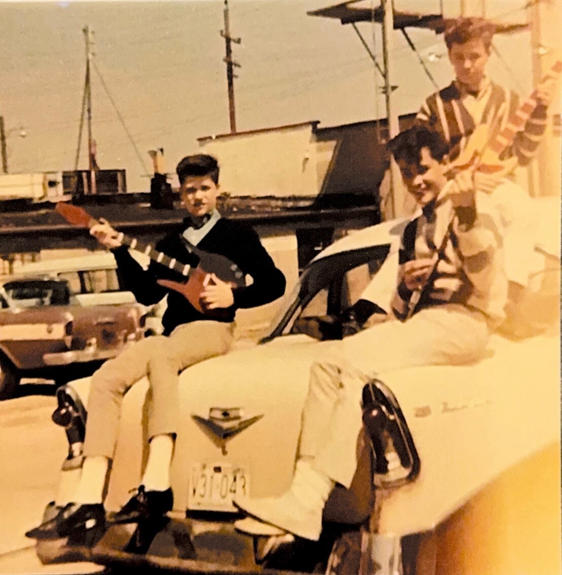 Three young men pose on a vintage car in a sunny street; one holds a red guitar-like instrument while the others pose nearby.