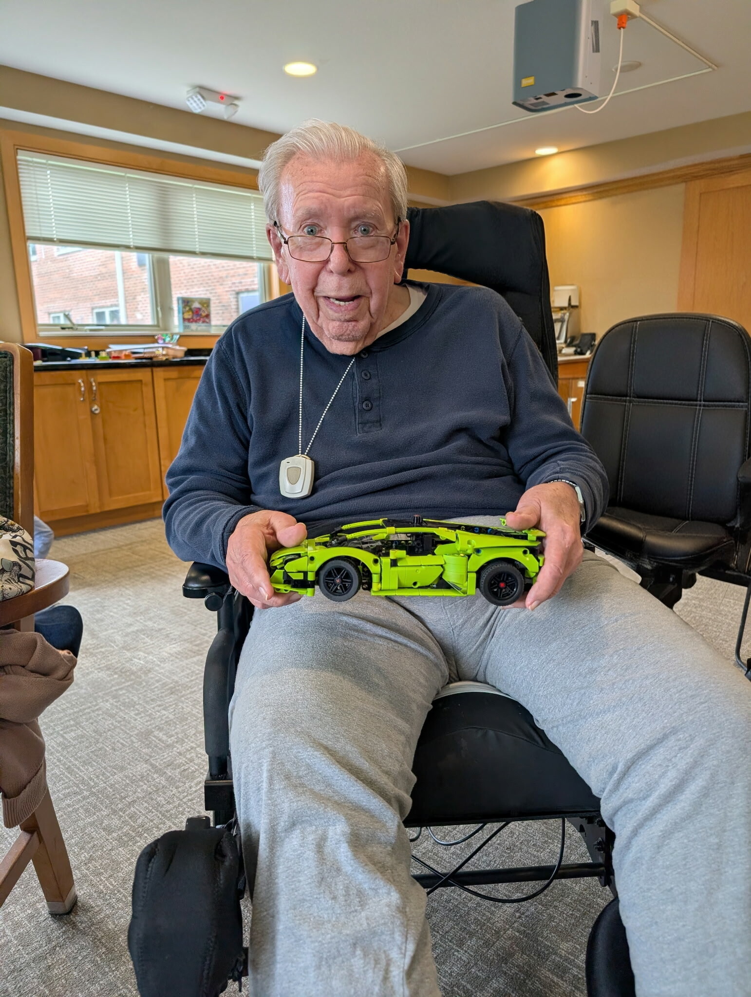 Older man in a wheelchair holds a bright green toy car model toward the camera in a sunny room.