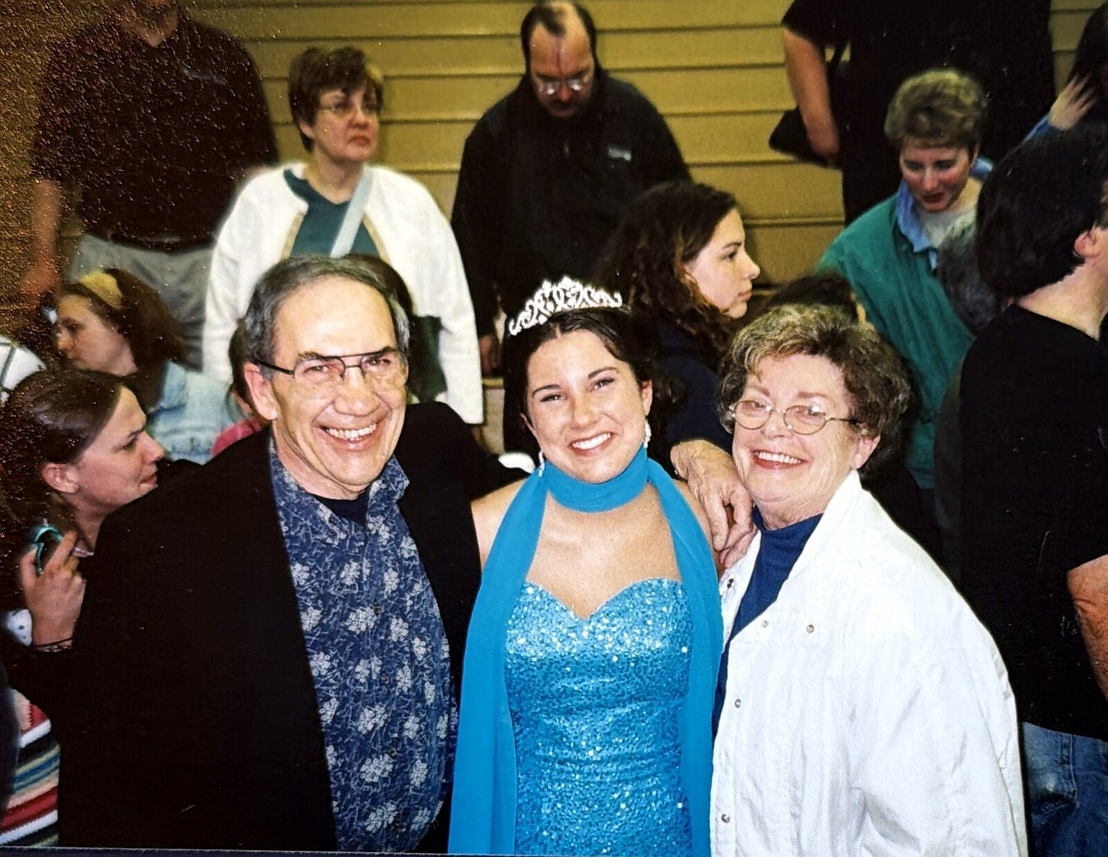 Young woman in a blue sequined gown and tiara posing with two older adults at a crowded event.