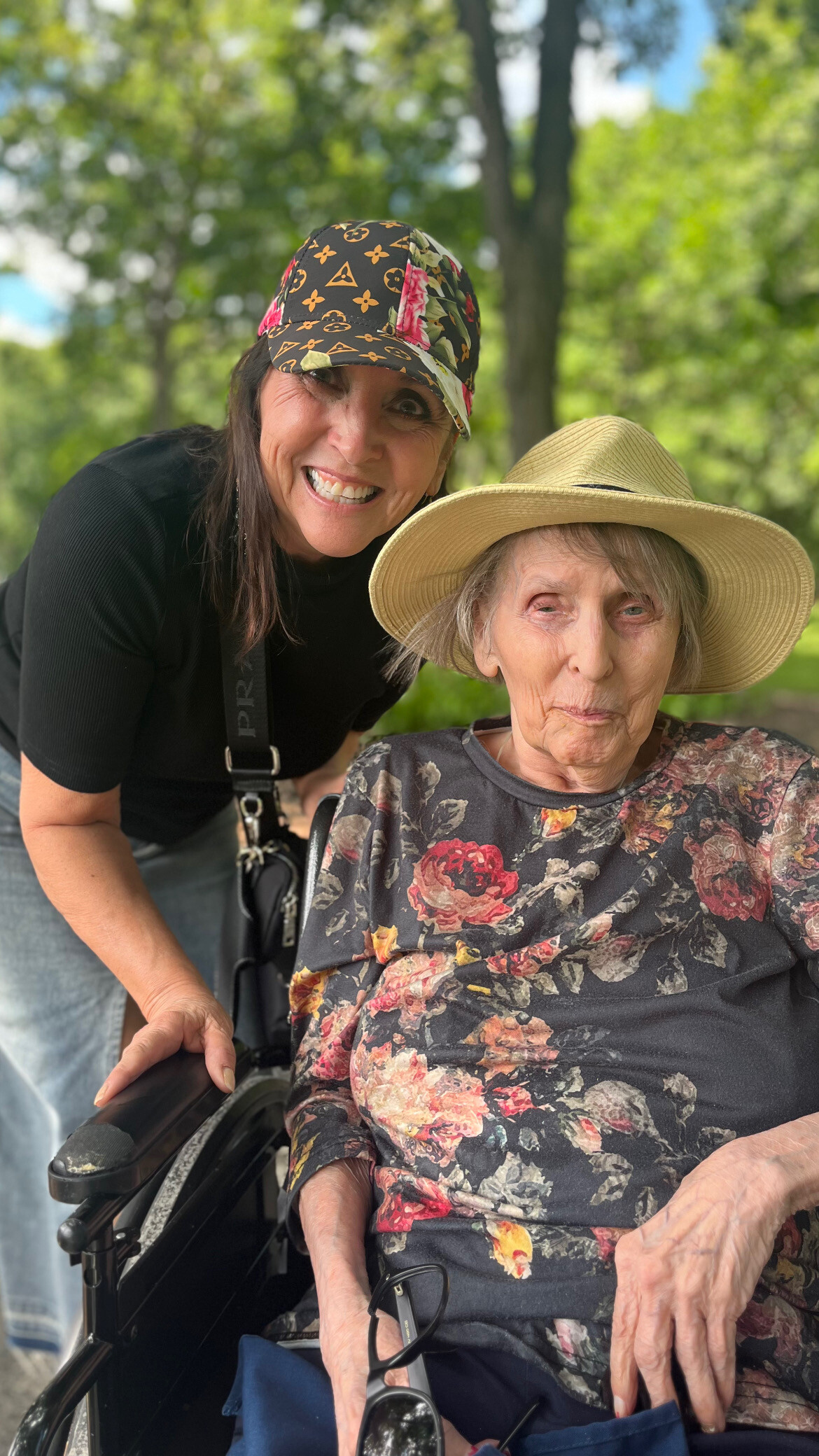 Two women outdoors: an older woman in a floral blouse sits in a wheelchair, while a younger woman leans in with a patterned cap, both smiling.