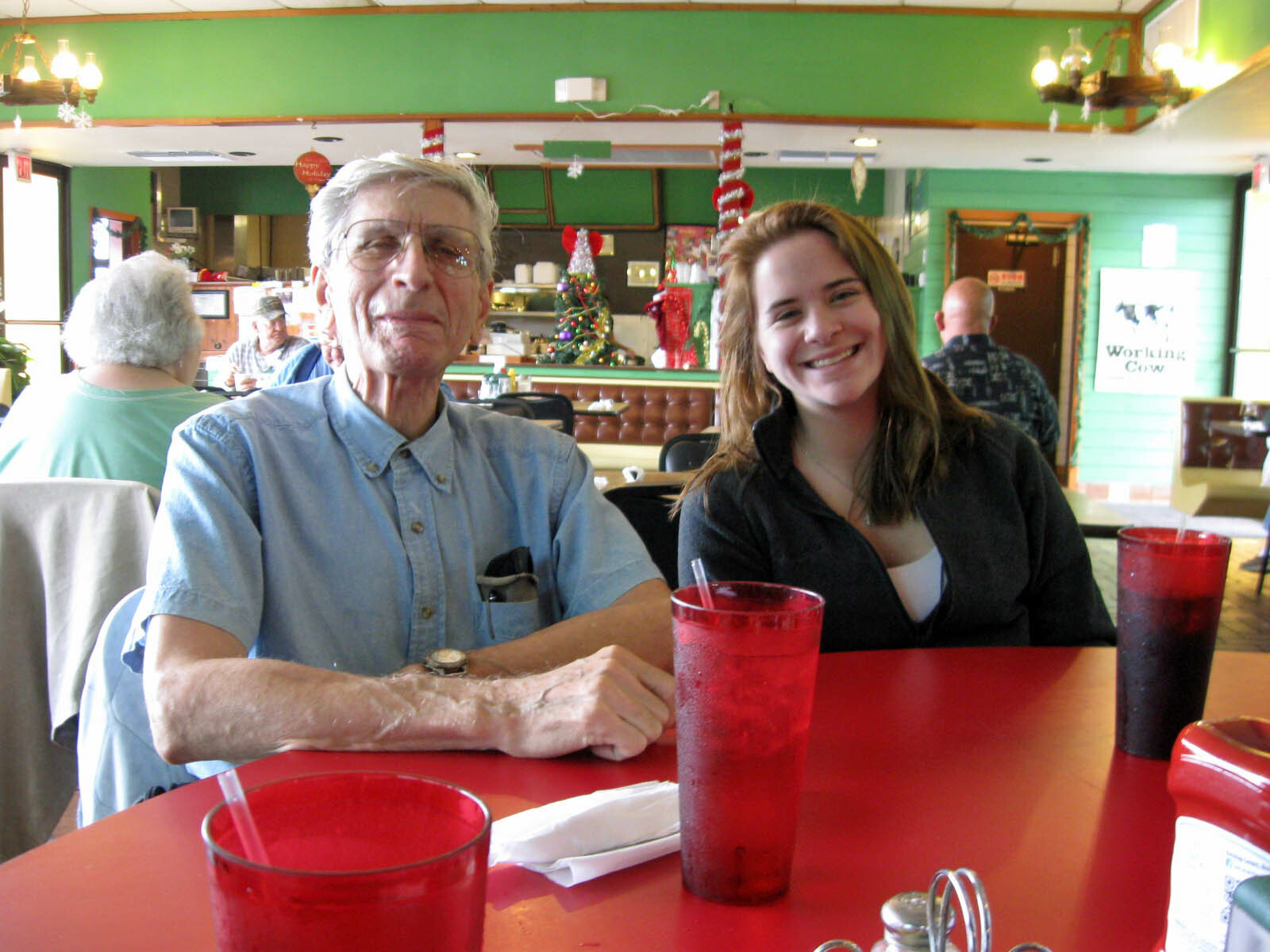 Older man in a blue shirt and a smiling young woman sit at a red table in a festive green-diner setting with Christmas decorations in the background.