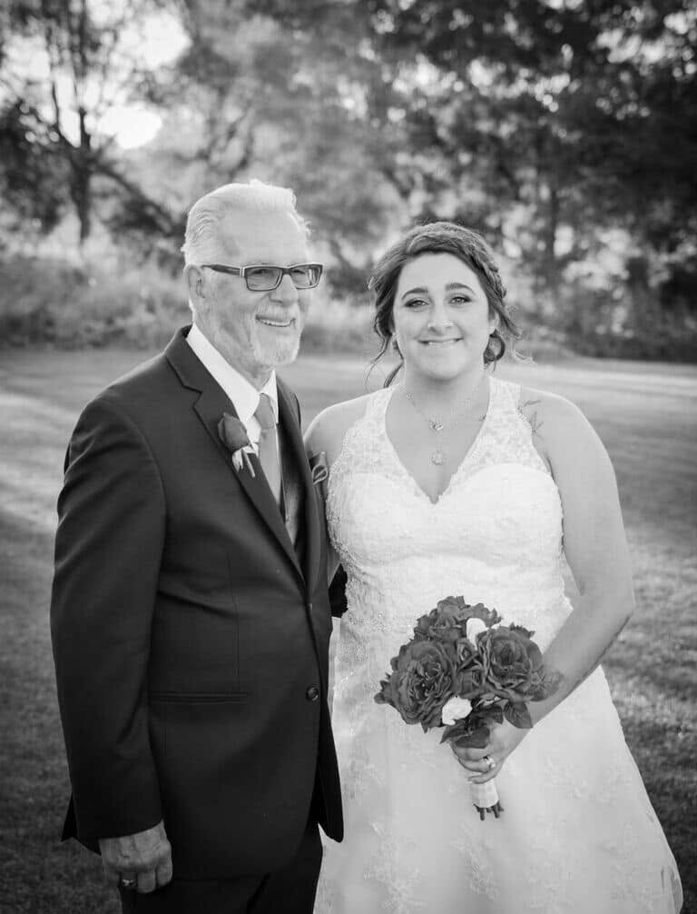 Bride in a lace wedding gown holds a bouquet, standing with an older man in a suit outdoors at a wedding venue.