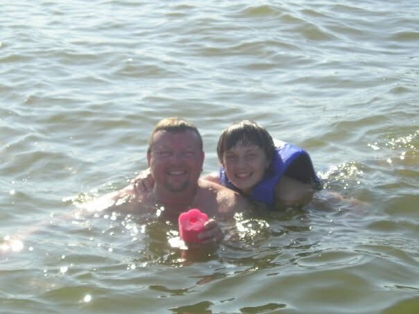 Man and child swimming in a lake, both smiling, wearing a blue life jacket, holding a pink toy.