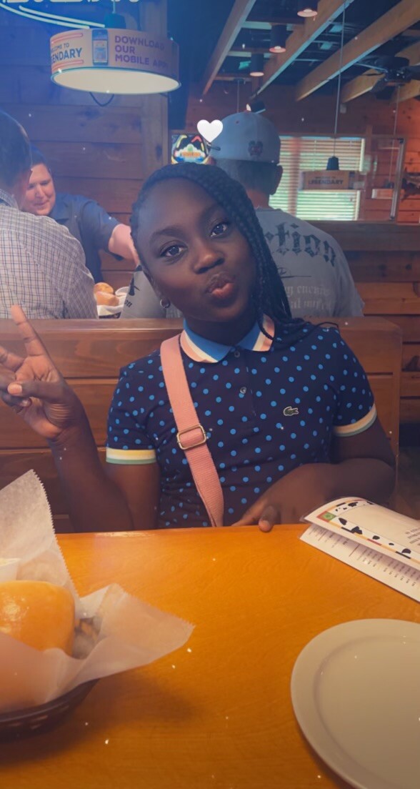 Girl at a restaurant table wearing a blue polka-dot polo and pink crossbody bag, making a peace sign and puckered lips toward the camera.