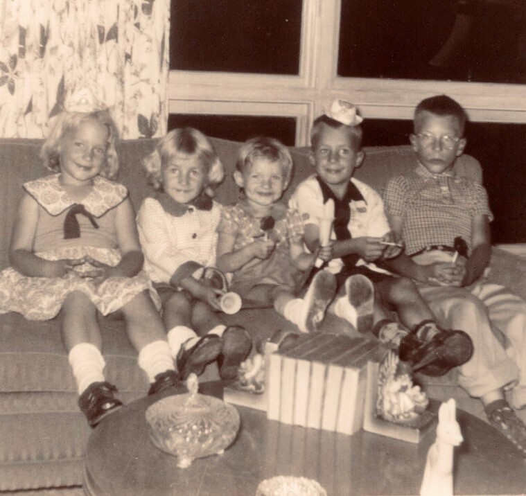 Six young children sit side by side on a couch, each holding small toys, in a vintage living room with a window and patterned curtains behind them.