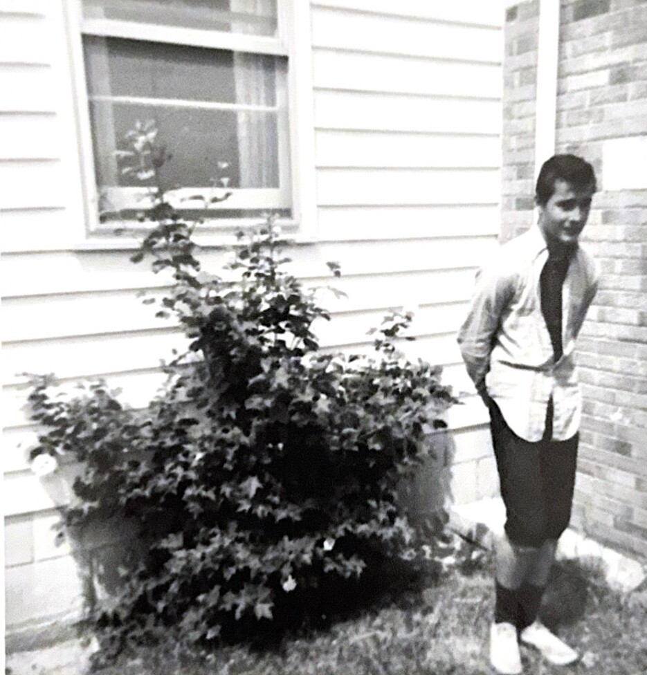 Black-and-white photo of a man in a shirt and tie standing beside a large shrub by a house.