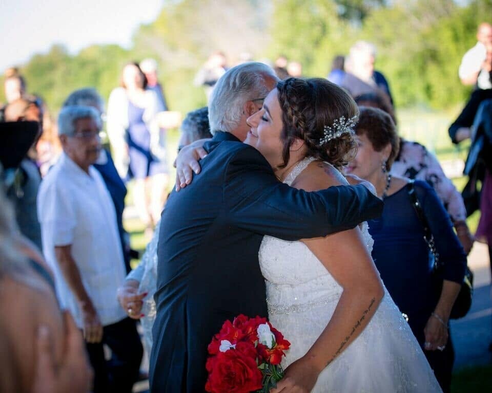 Bride in a white wedding gown hugs an older man outdoors; a red bouquet rests near her waist in the foreground.