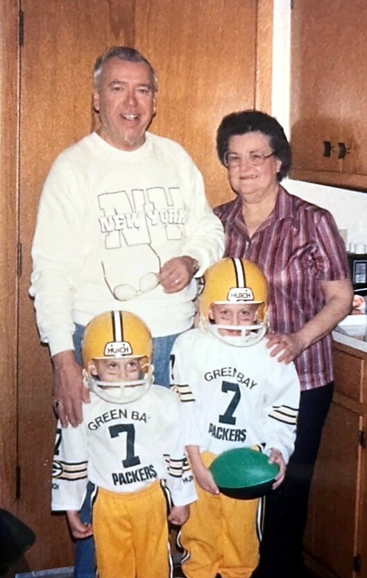 Family of four posing in a kitchen; two young boys in yellow Packers football uniforms and helmets stand in front, adults behind them.