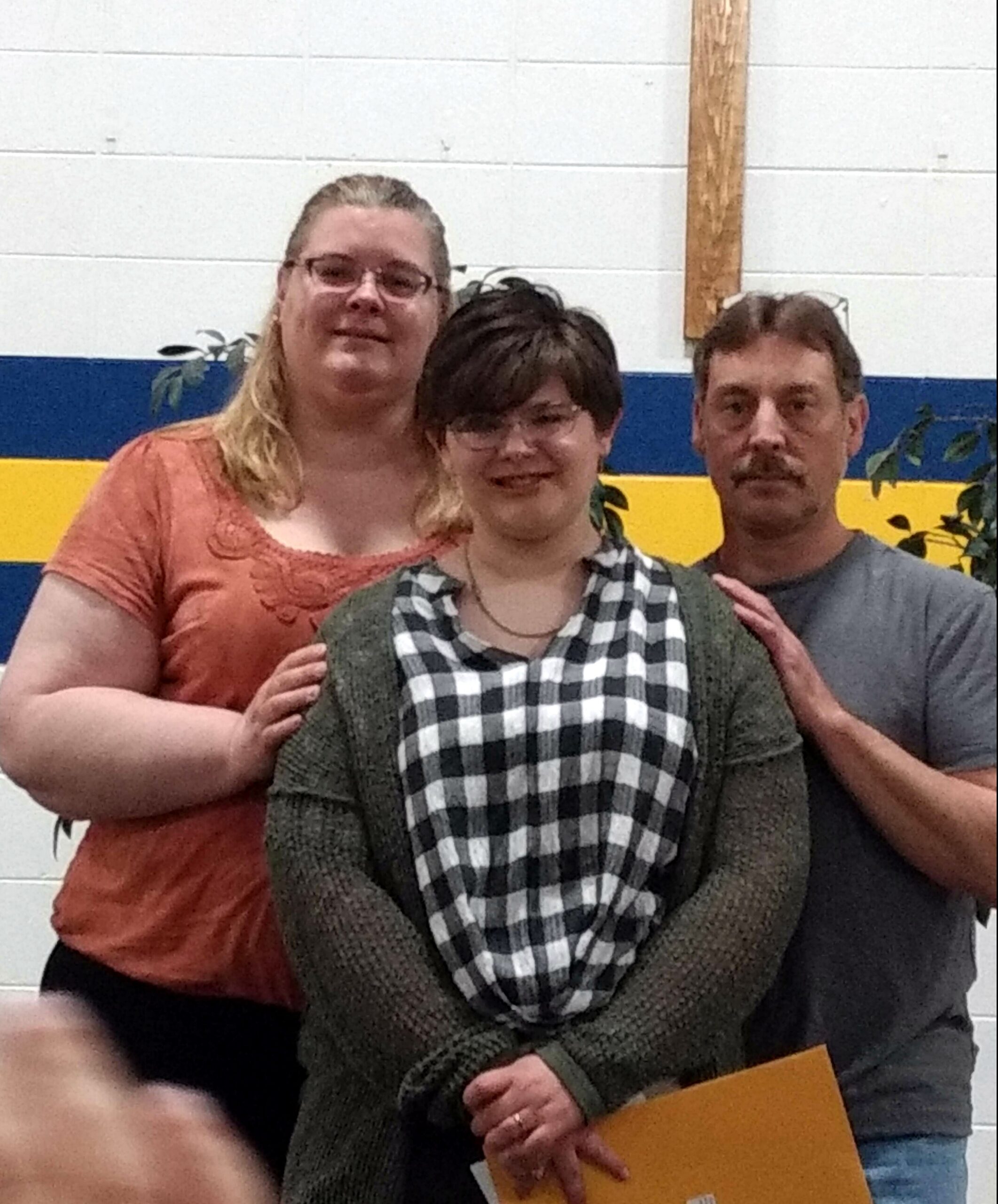 Three people posing indoors in front of a blue and yellow wall; a woman in an orange top on the left, a person in a plaid shirt in the center, and a man in a gray shirt on the right.