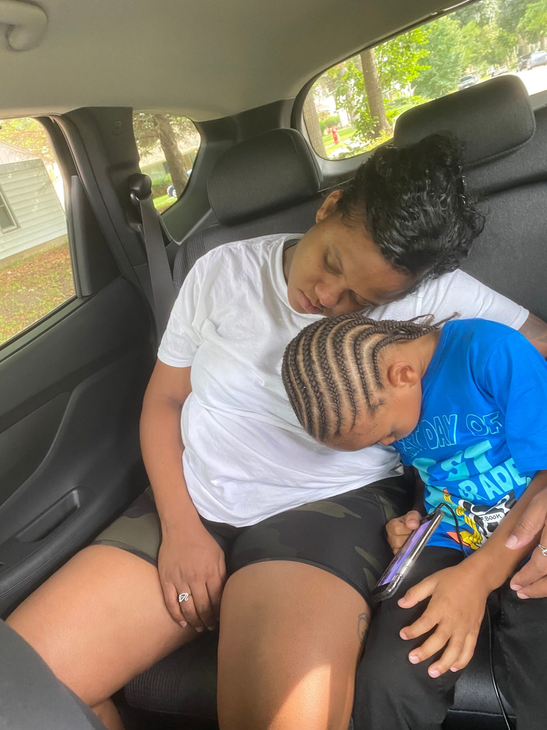 Two people sleep in the back seat of a parked car: an adult in a white shirt leaning toward a child with braided hair wearing a blue shirt.