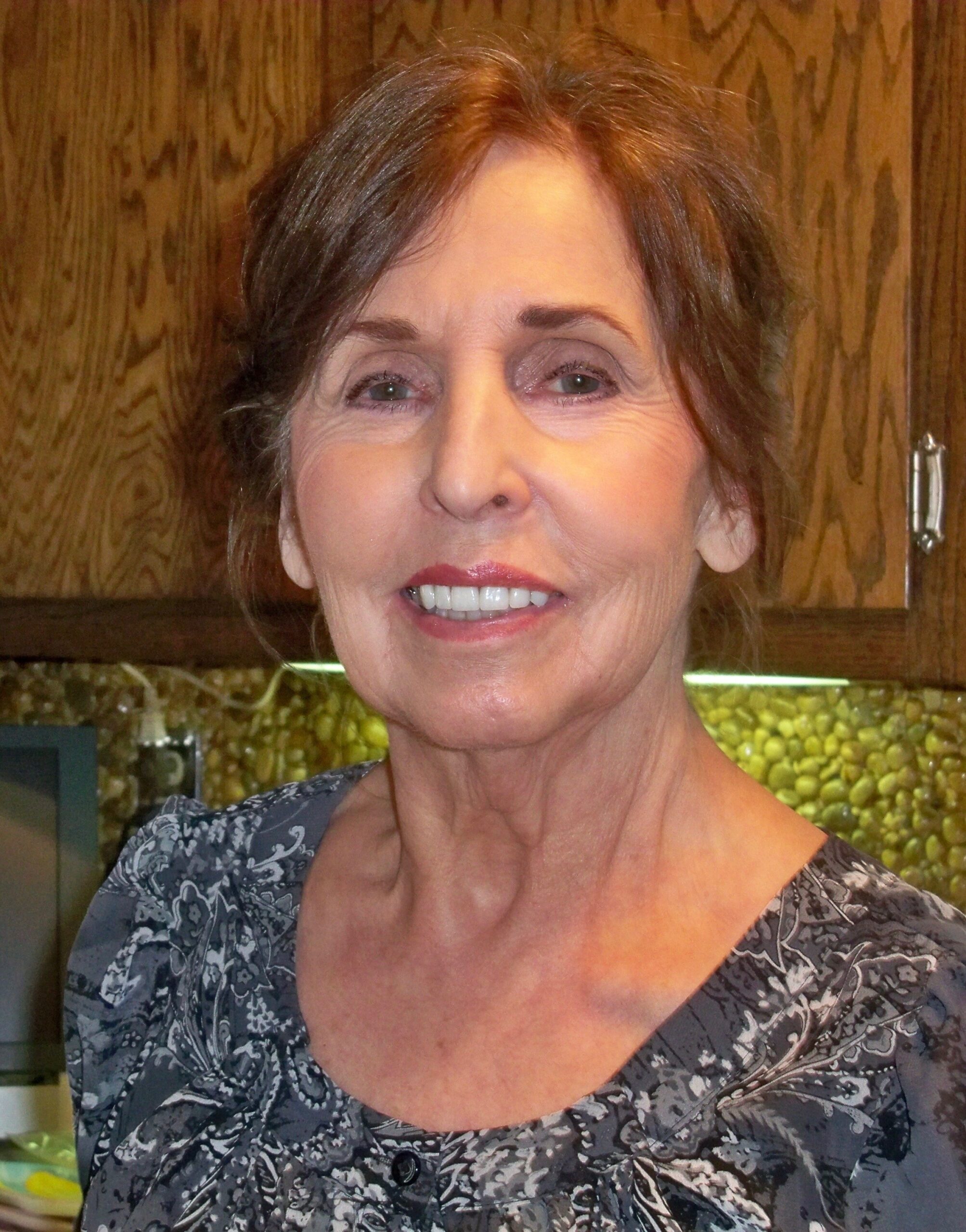 Close-up of a smiling woman with brown hair wearing a gray floral-patterned blouse, wooden cabinets in the background (kitchen).