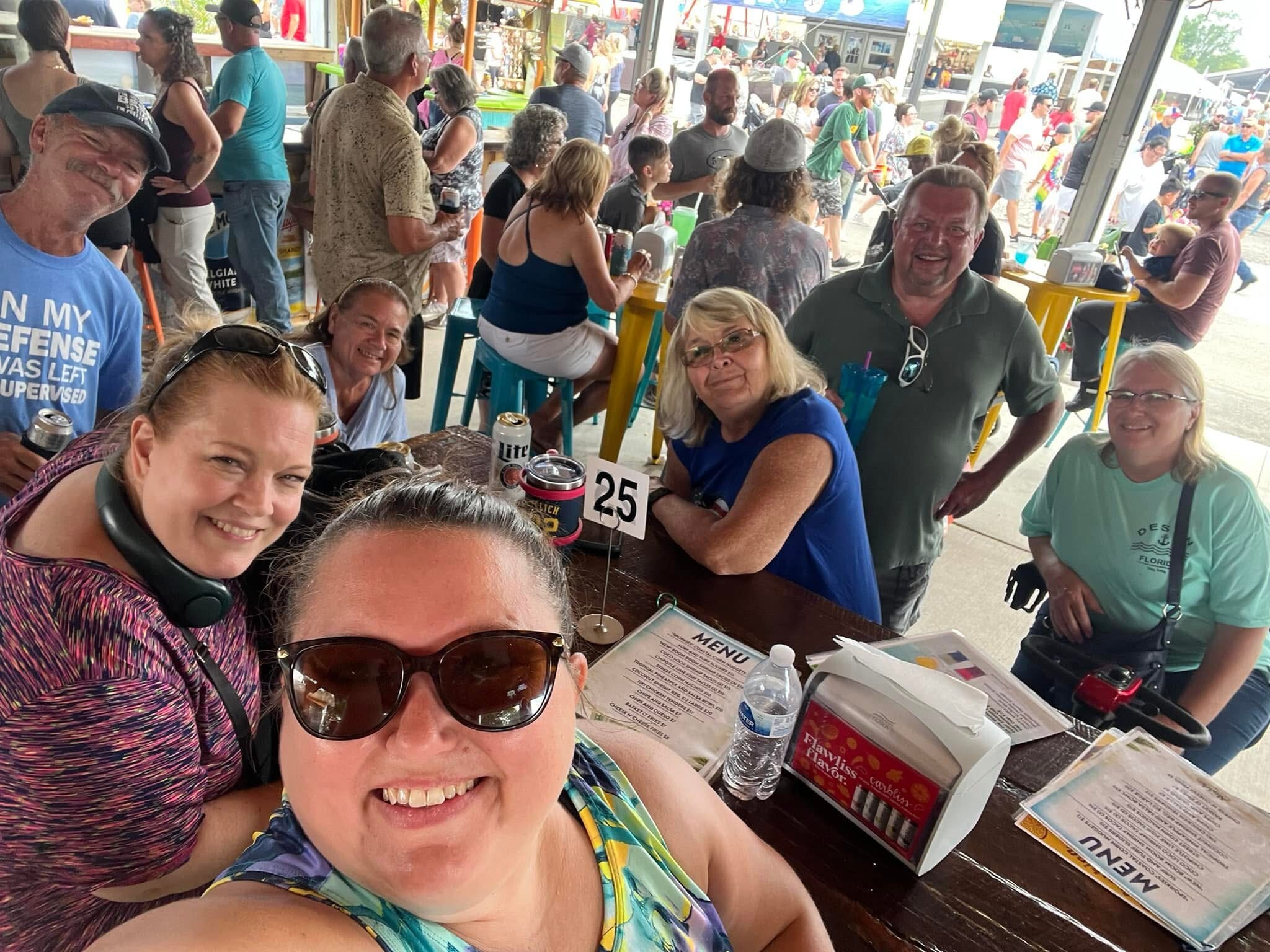 Group selfie at a crowded outdoor bar/event, smiling people around a table with drinks and menus in view.