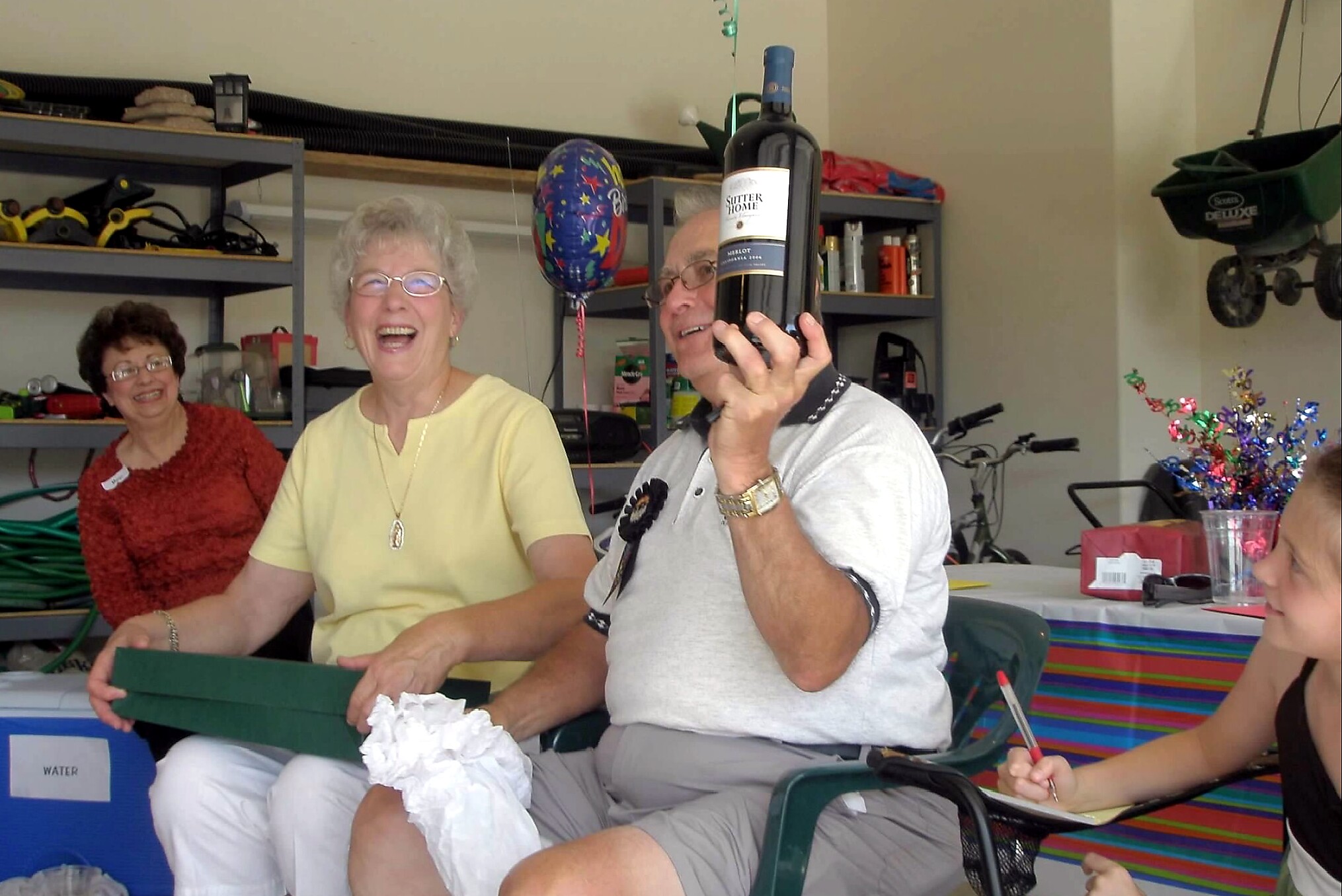 A group of smiling seniors at a party in a workshop; man holds up a wine bottle beside a starry balloon while others chat and laugh.