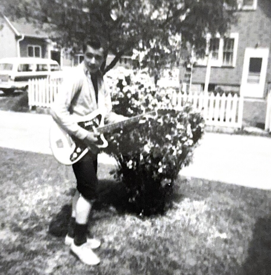 Man standing in a front yard playing a white electric guitar, with a bush and white picket fence behind him in a suburban street (black-and-white photo).