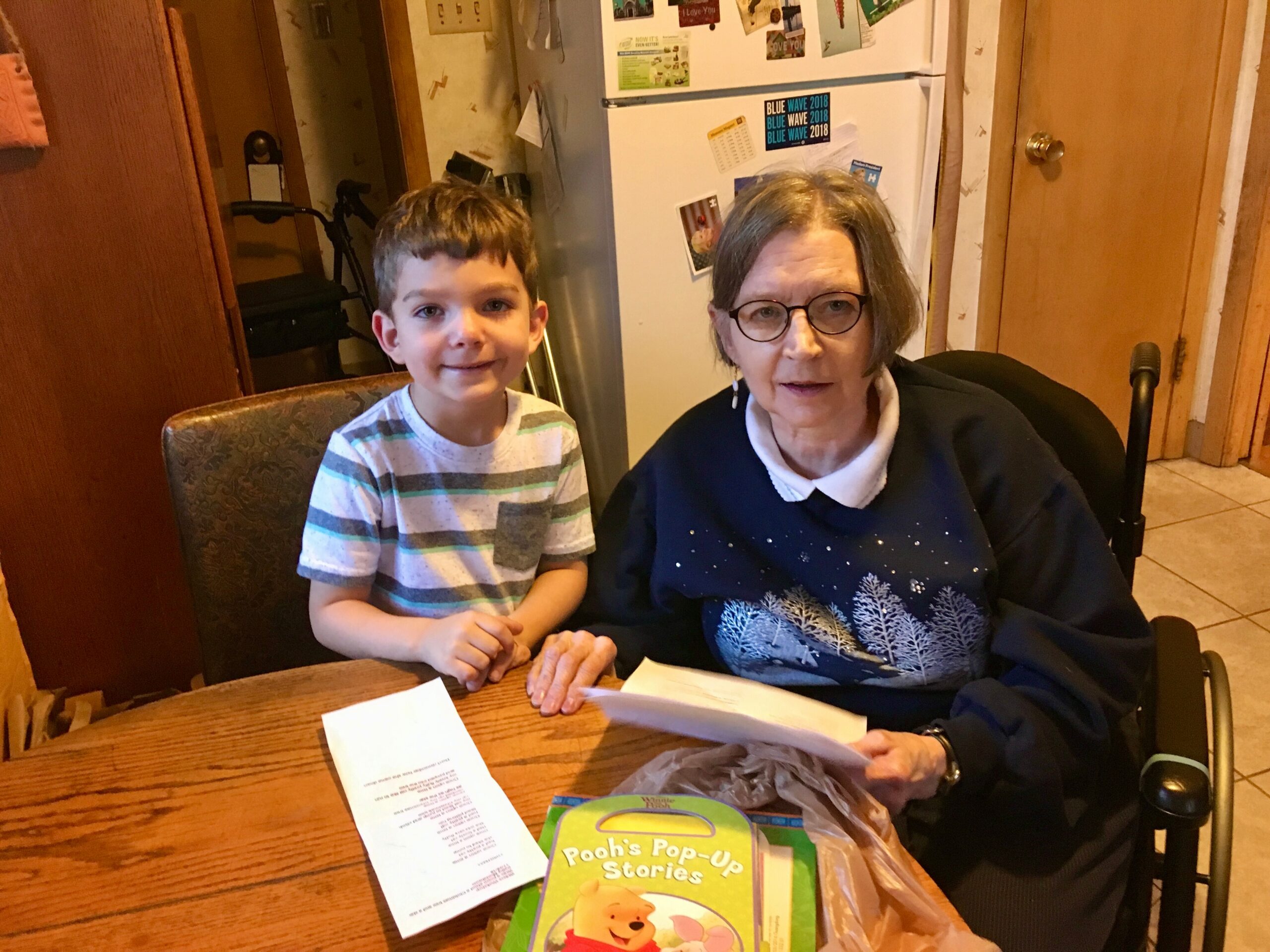 A smiling young boy and an older woman with glasses sit together at a wooden table, looking at papers on the table with a Pooh's Pop-Up Stories book visible in the foreground.