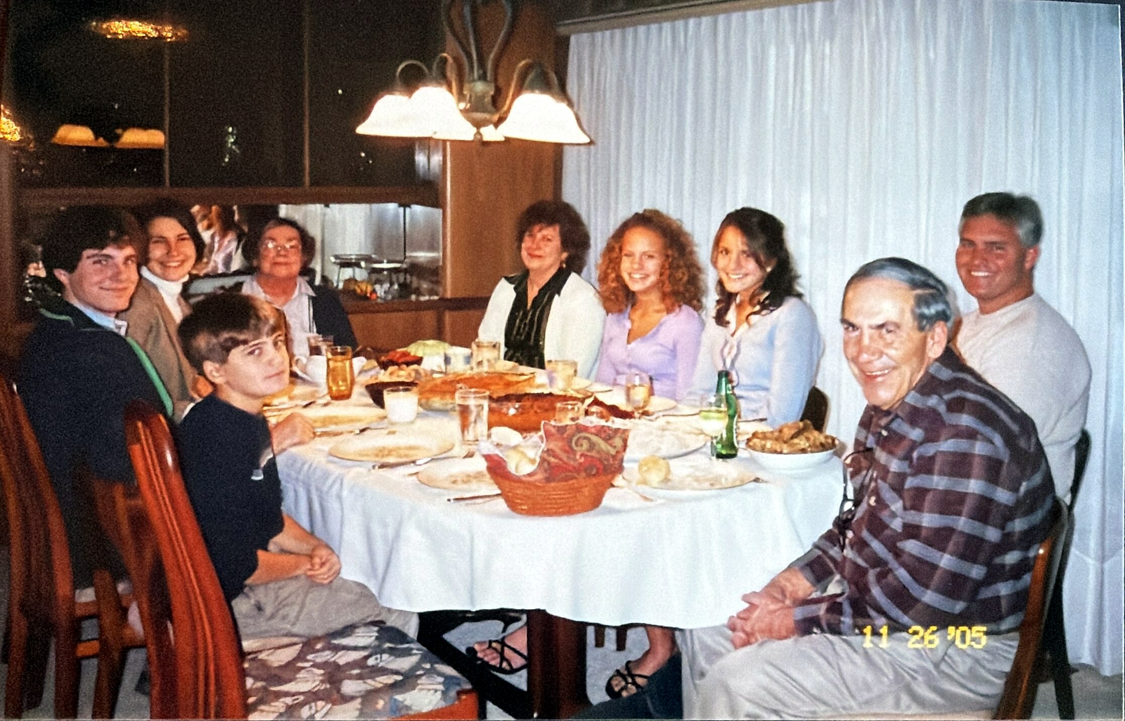 Group of nine people at a long dinner table in a restaurant, smiling, with bread baskets and plates; date stamp 11/26/05 visible in lower right.