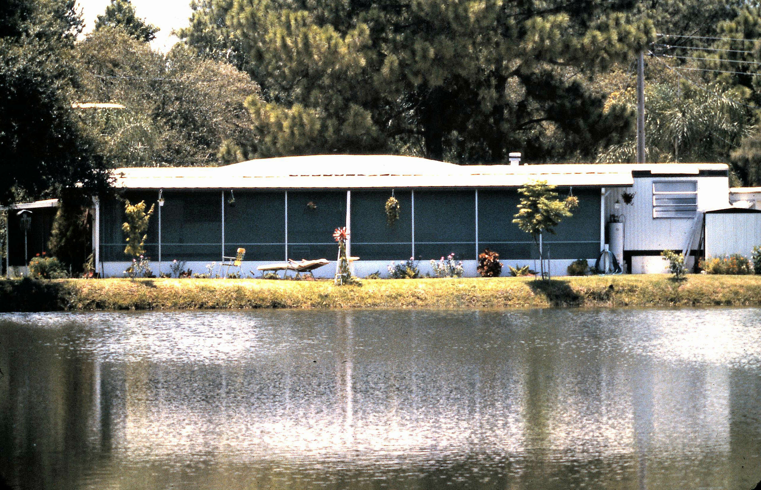 One-story house with a long screened porch by a body of water, surrounded by trees and a small garden in the yard.