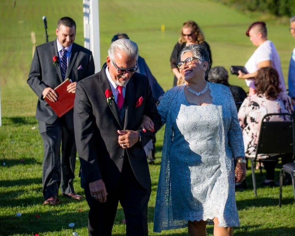 Senior couple walking arm-in-arm at an outdoor wedding ceremony, the man in a dark suit with a red rose boutonniere and the woman in a blue lace dress, smiling as they stroll.