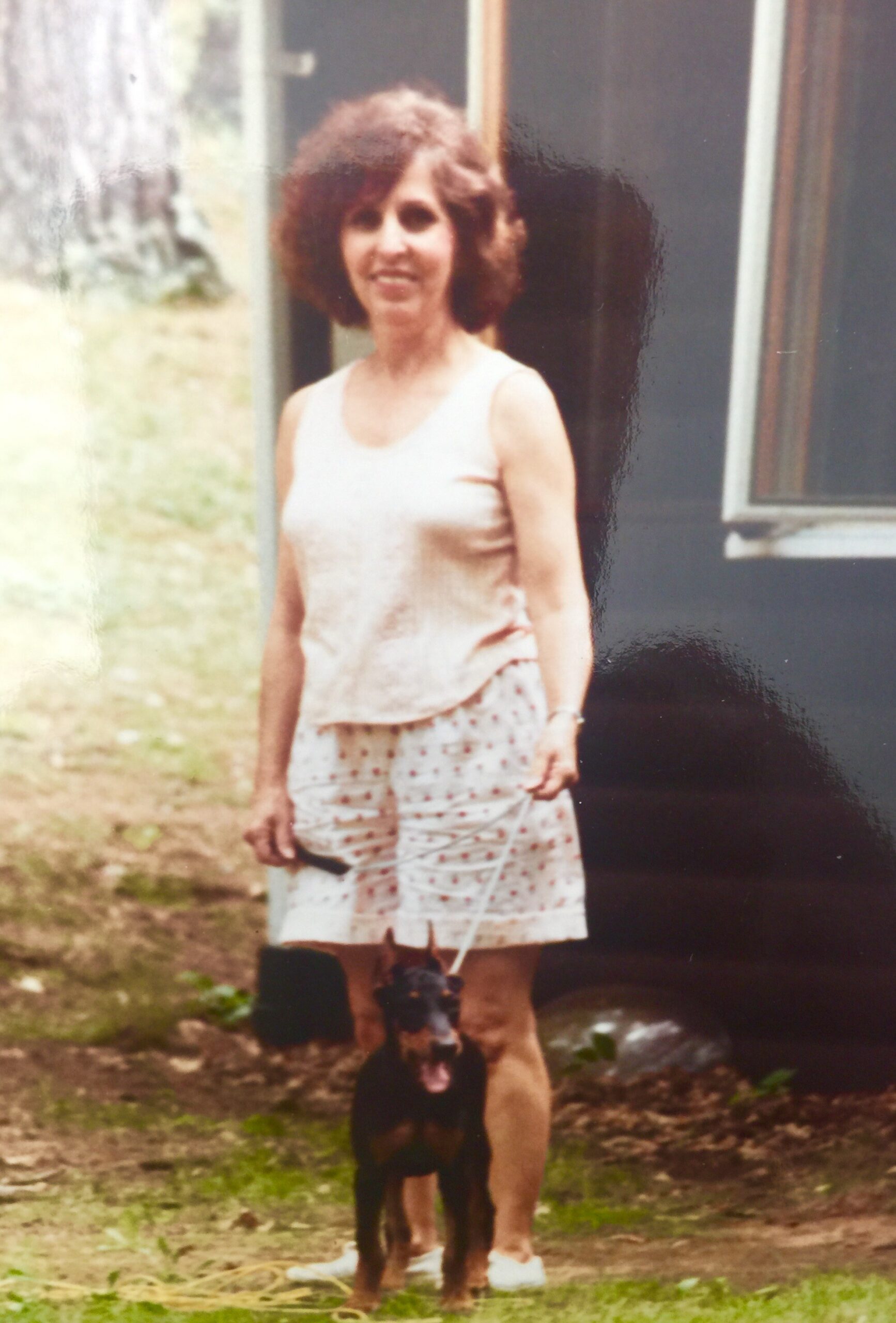 Woman in a light sleeveless top and floral shorts stands outside with a small black dog by her feet.