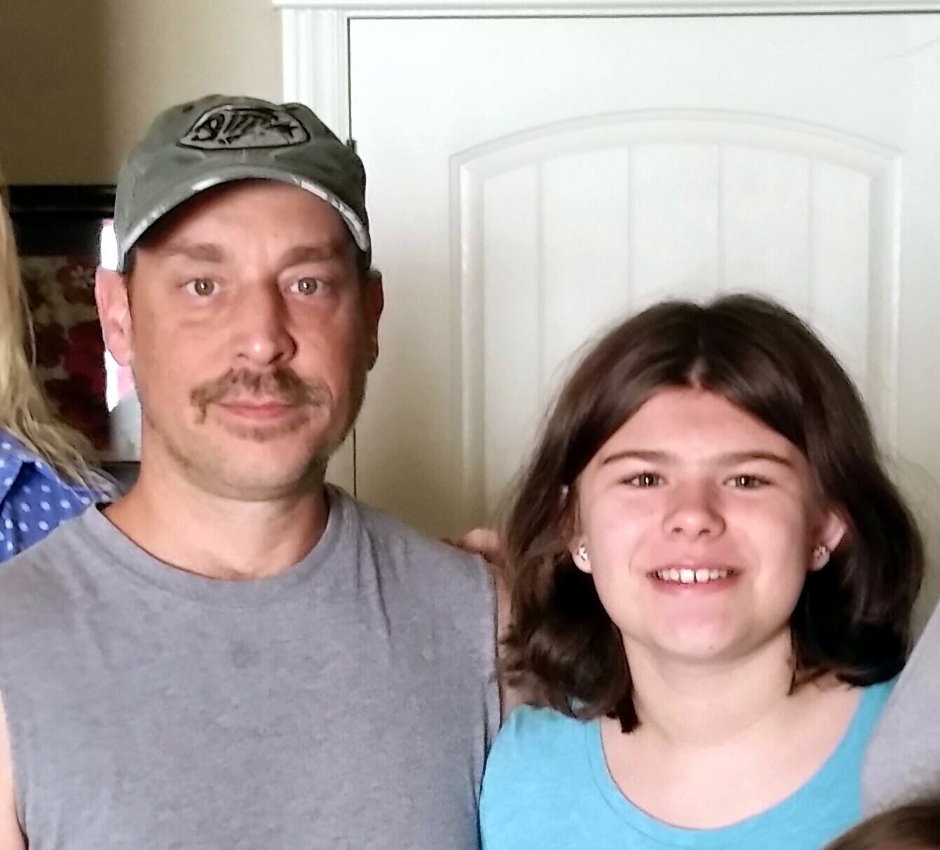 Man in gray shirt and cap stands next to a smiling girl in a turquoise top indoors.