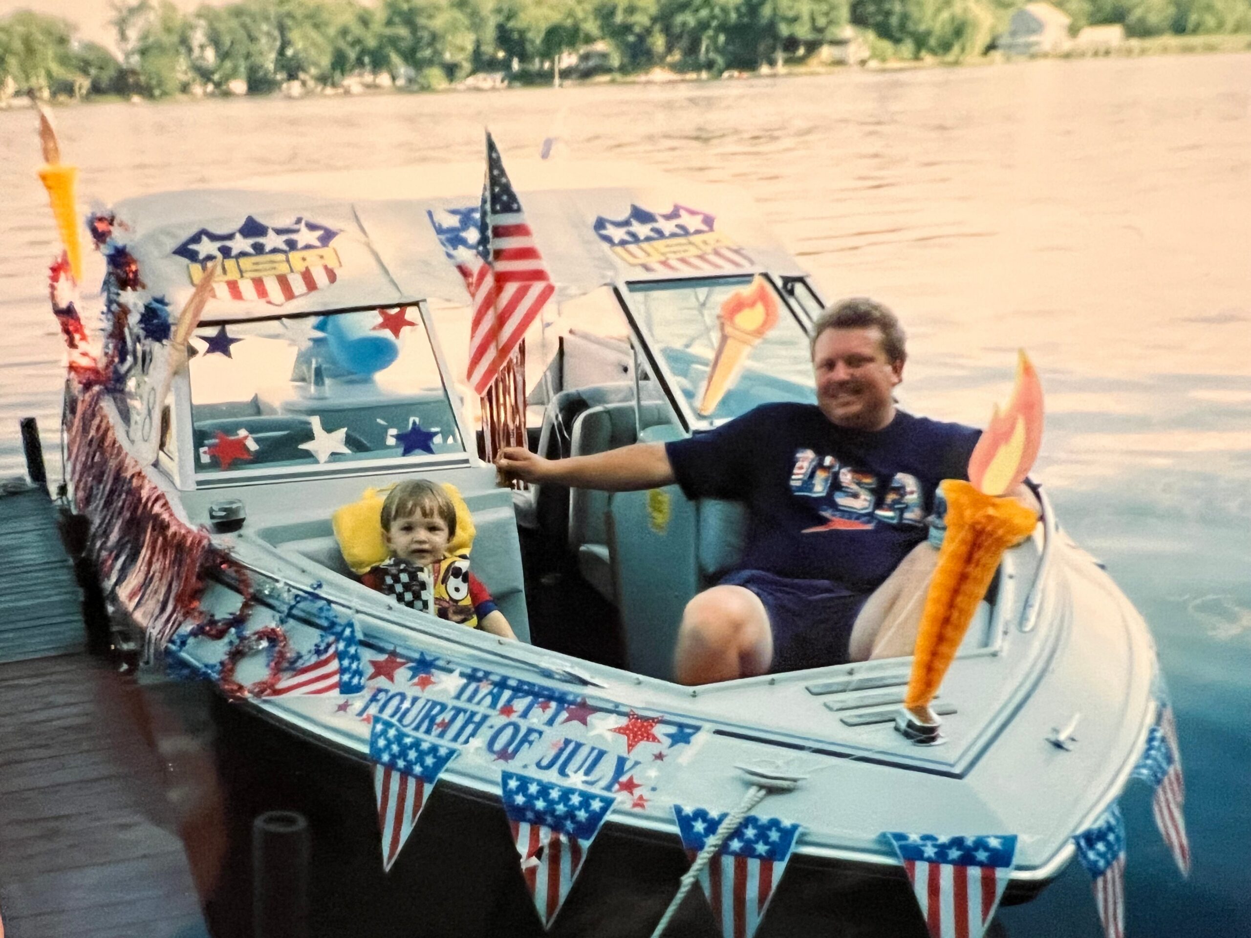 Man and child in a small boat decorated with American flags and Fourth of July bunting on a lake surface.