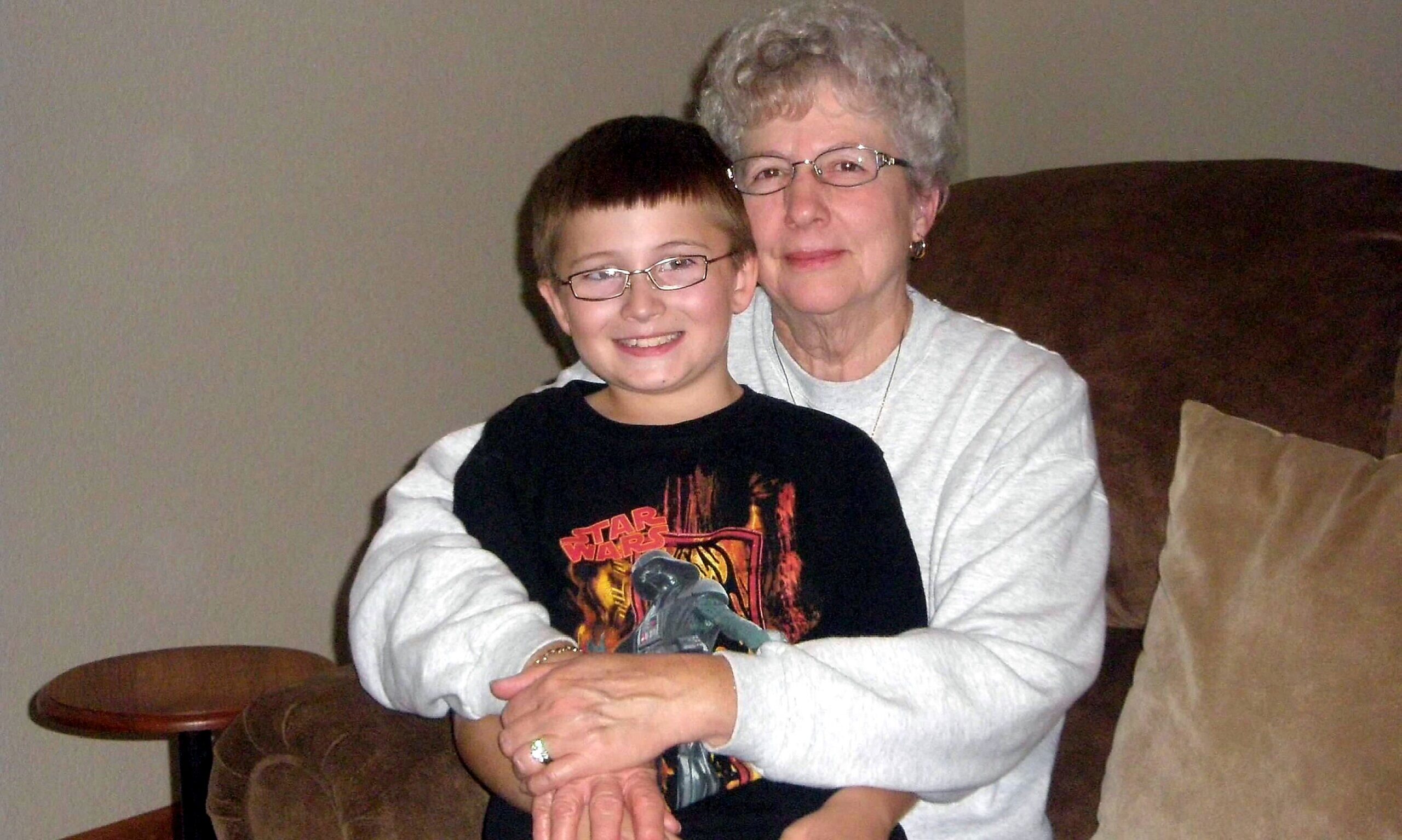 Smiling boy with glasses sits with his grandmother on a brown couch in a living room, both facing the camera.