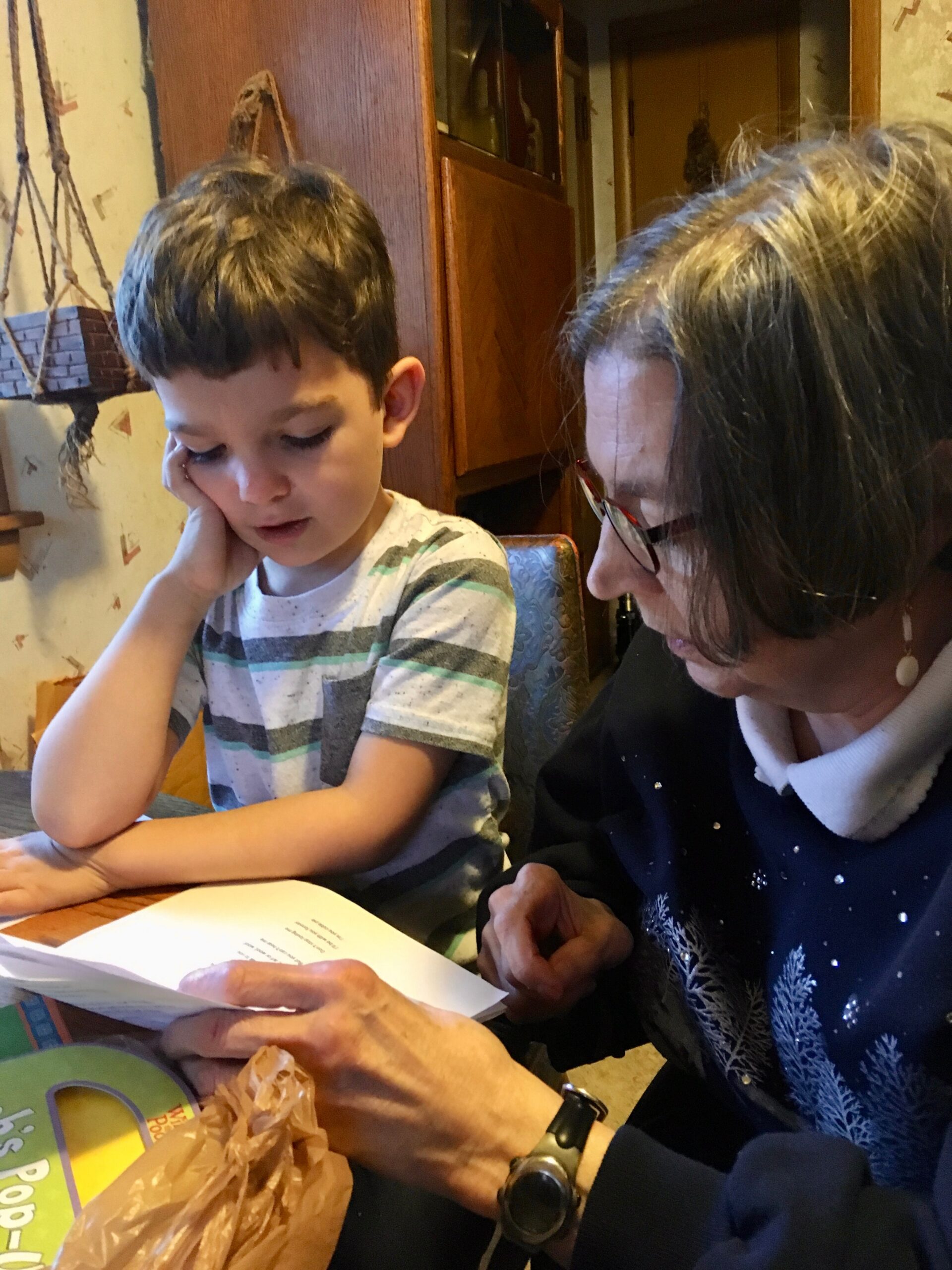 Young boy resting his head on his hand while reading a paper at a table with an adult woman beside him looking on thoughtfully,