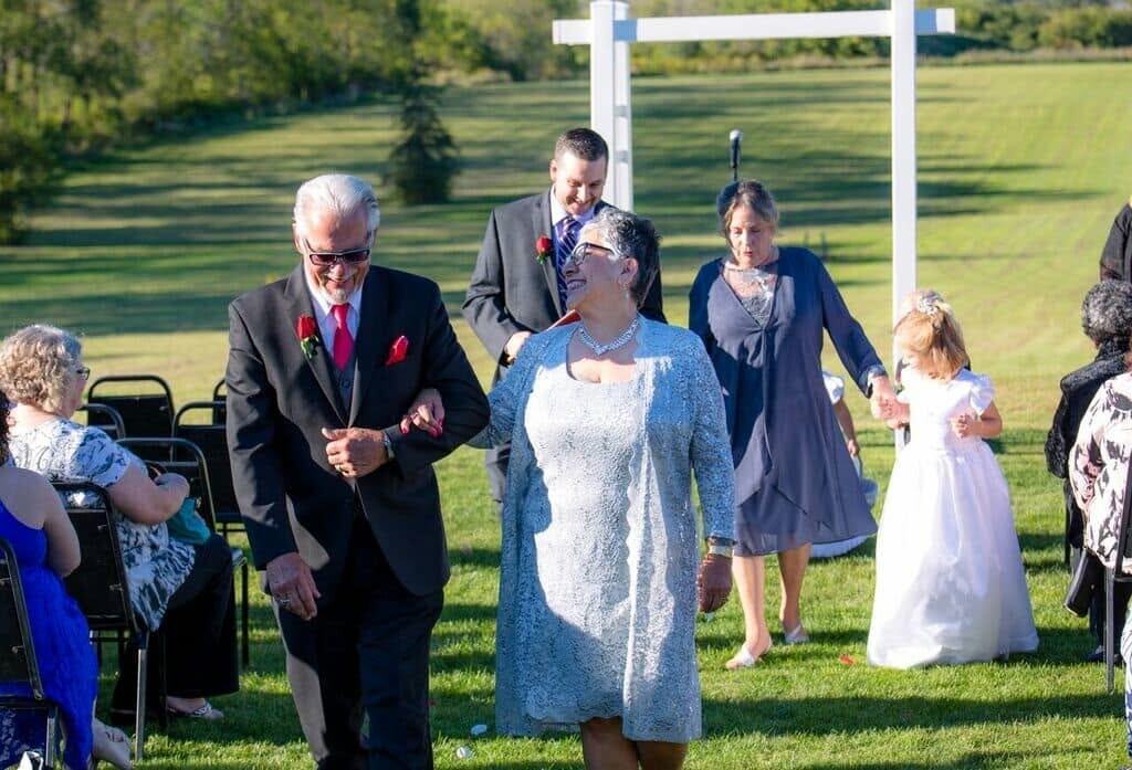 Elderly man in a black suit with red boutonniere and a woman in a blue lace dress walk arm in arm down an outdoor grassy aisle, smiling as wedding guests sit nearby.
