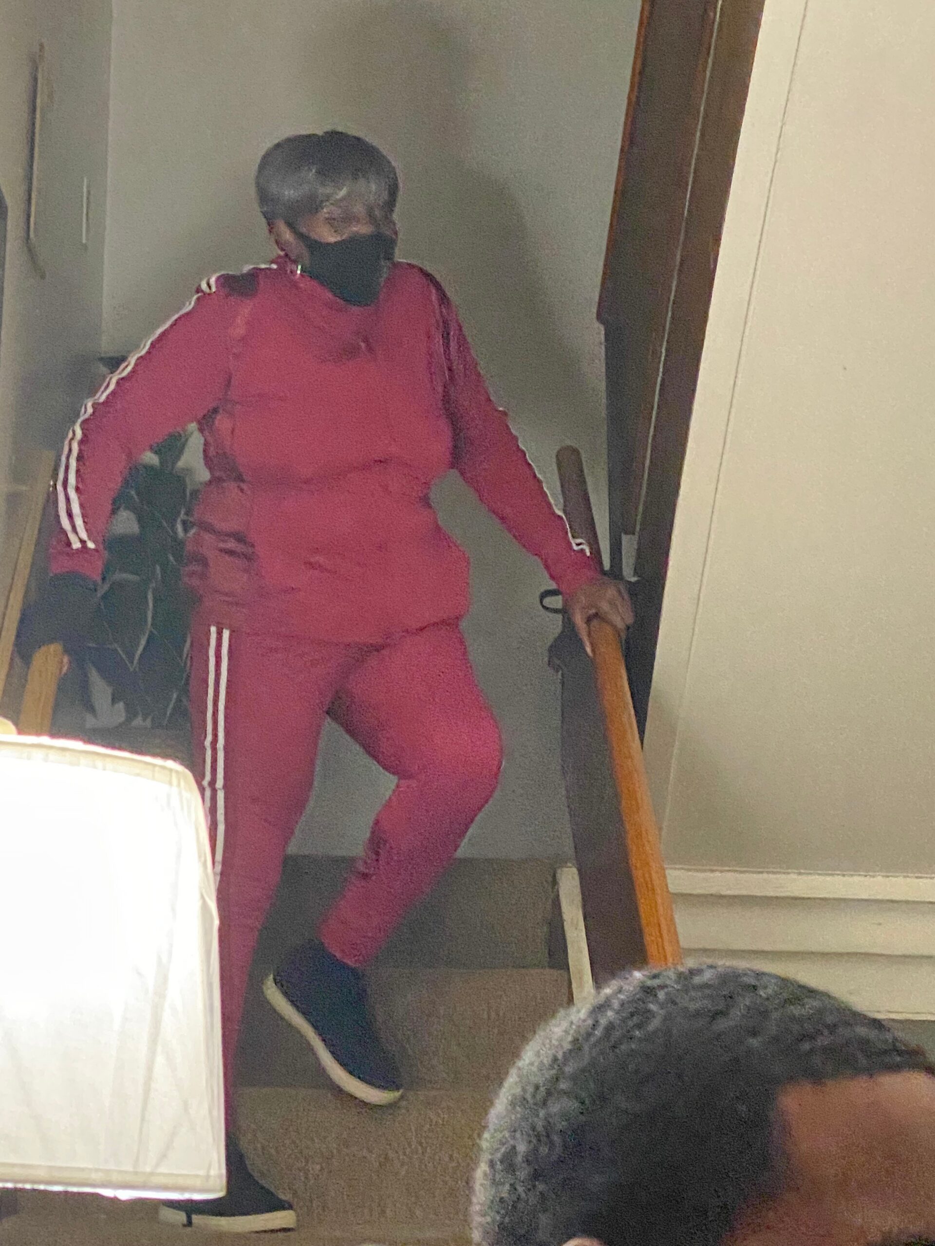 Person wearing a red tracksuit with white stripes standing on a staircase indoors, holding the railing and wearing a black face mask.