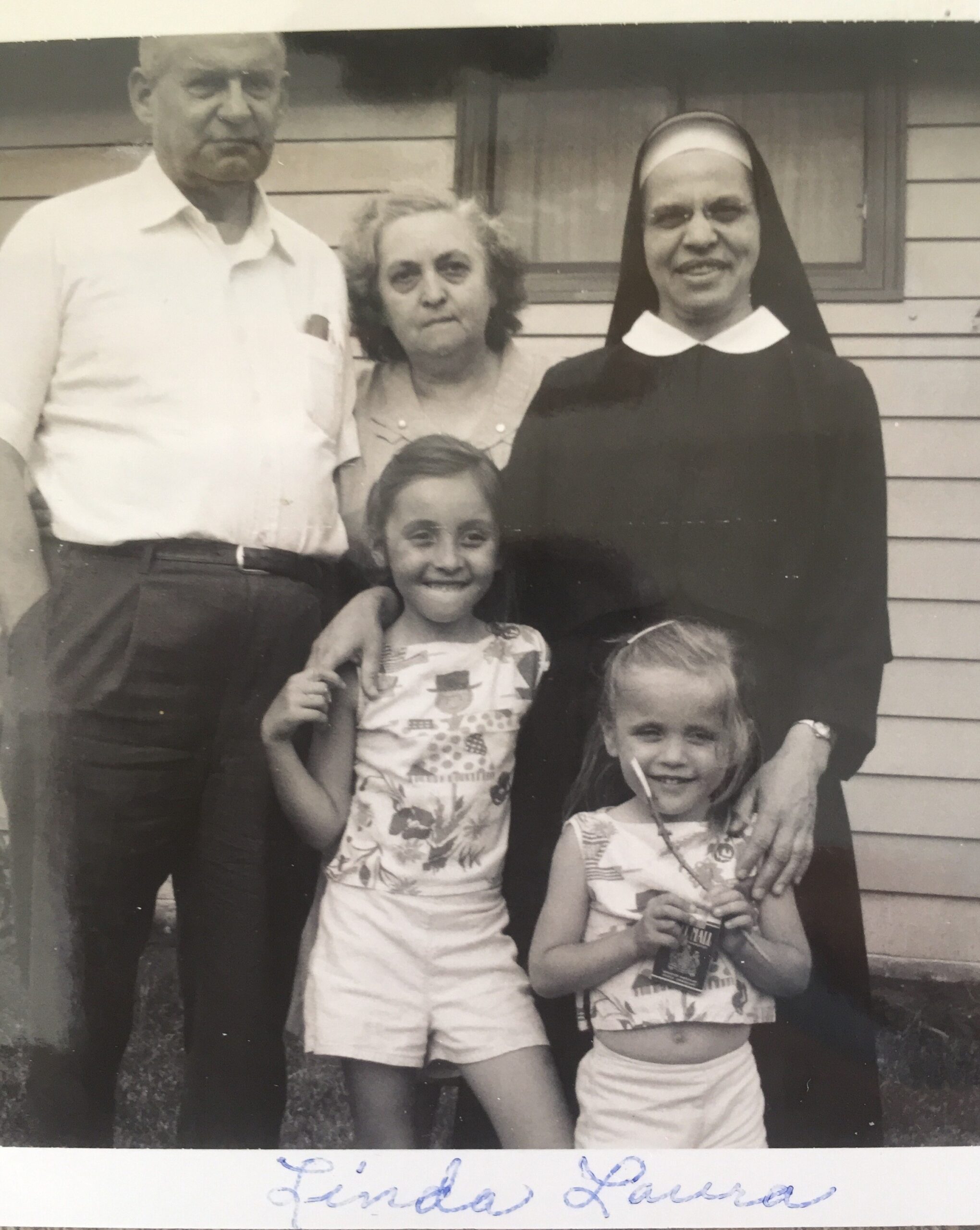 Group portrait of an elderly man, two young girls, and a smiling nun in front of a house.