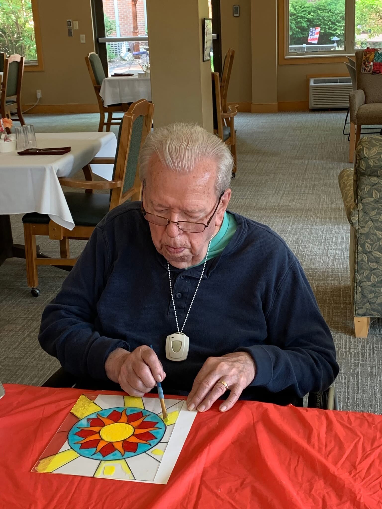 Elderly person seated at a table, painting a bright sunburst design on paper under a red tablecloth.