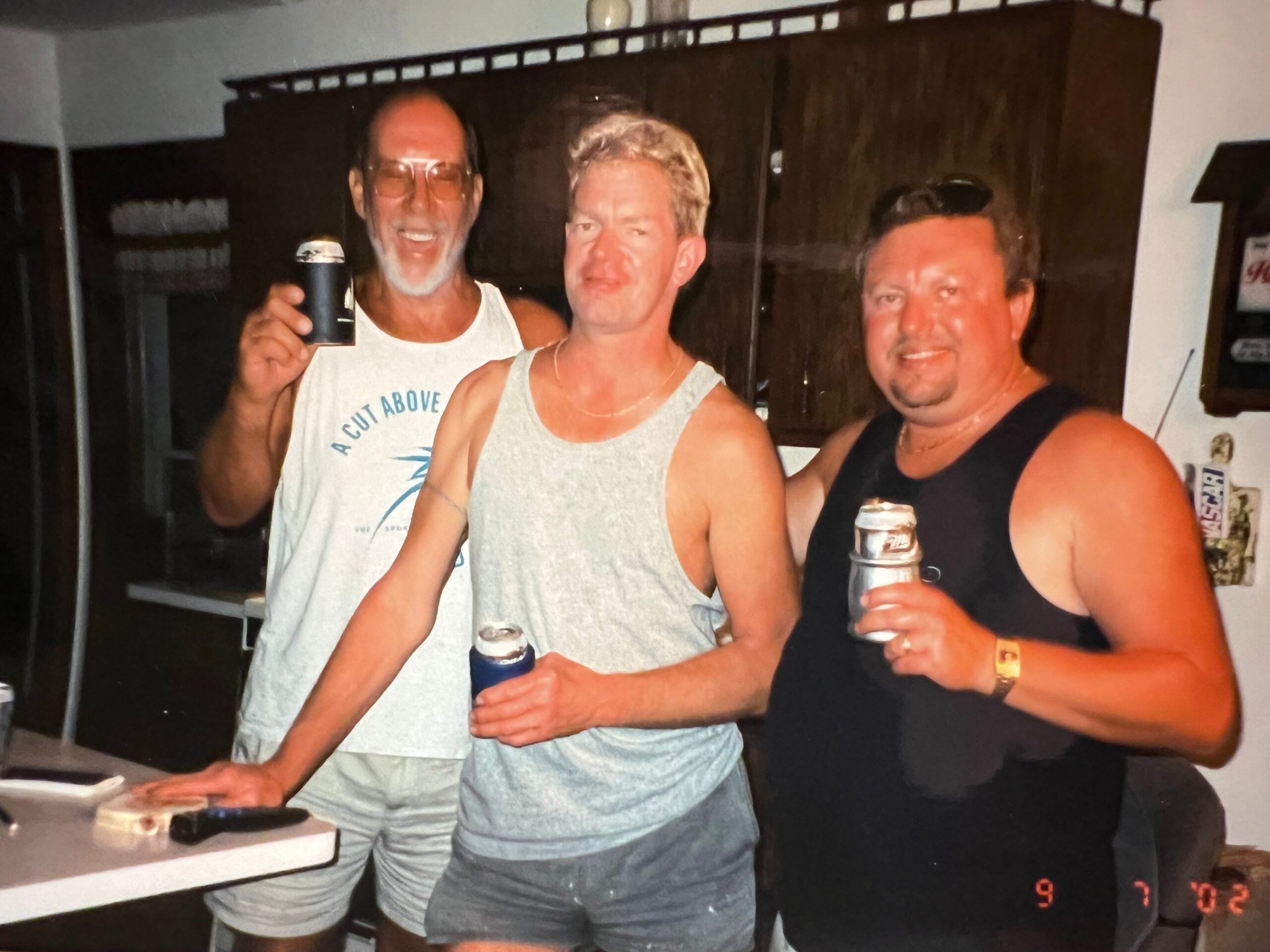 Three men in casual sleeveless tops smiling and posing with drinks at a social gathering indoors.