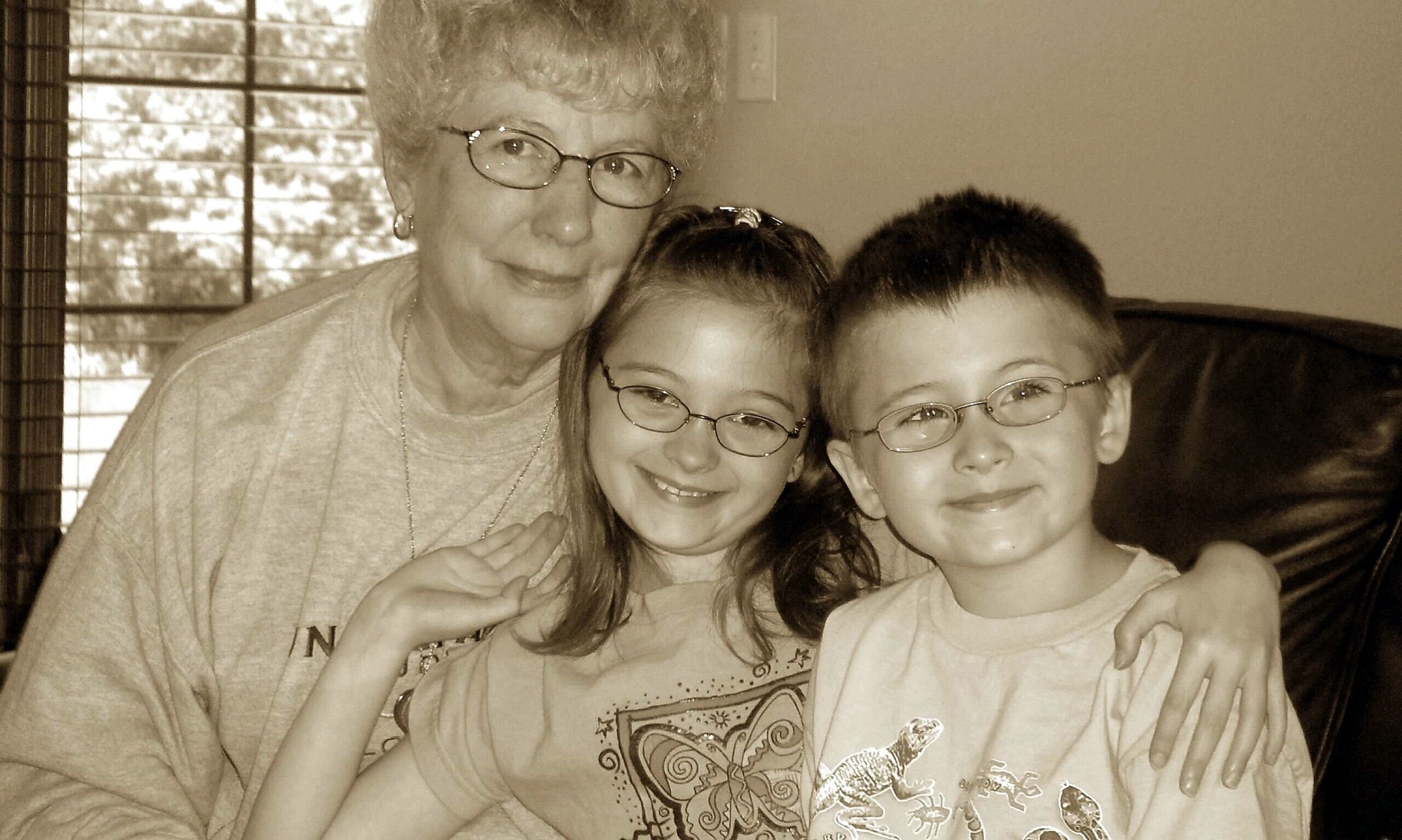 Grandmother wearing glasses hugs a smiling girl and boy indoors, all three posing for a family photo.