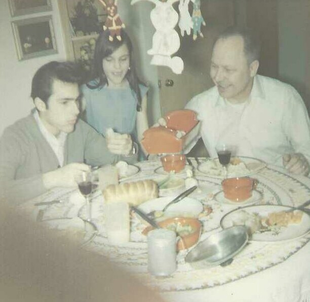 A family of three sits around a round dining table with bread, cups, and a red serving dish during a meal, smiling at each other.