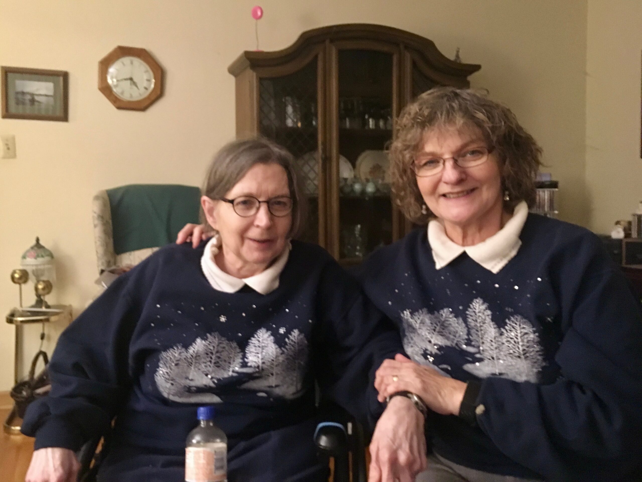 Two smiling older women sit together in a living room, wearing matching navy sweaters with white tree patterns behind them is a wooden china cabinet.
