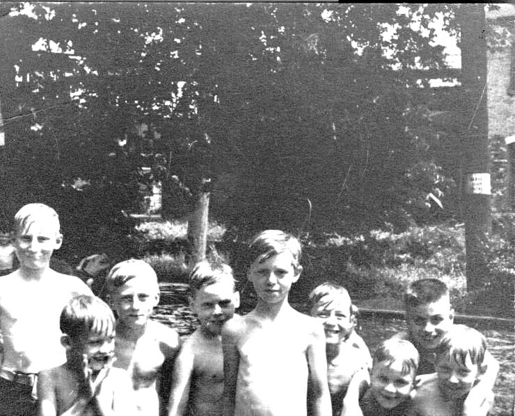 Eight shirtless boys pose together beside a small pond in a sunny park.