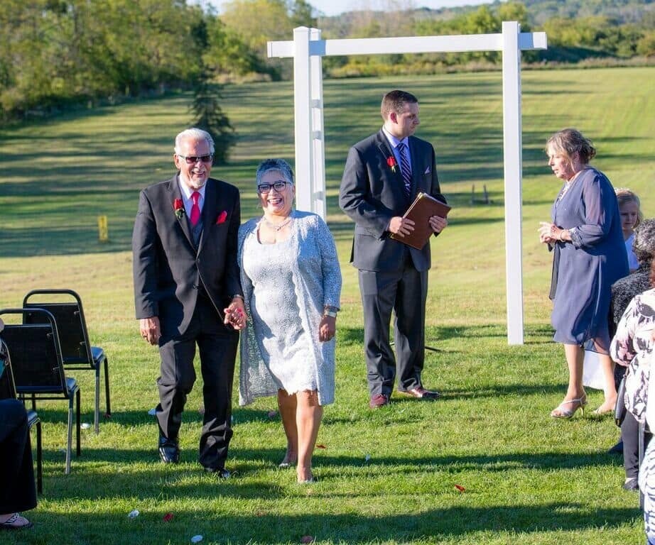 An elderly couple walking hand in hand down an outdoor grassy aisle at a wedding, guests seated nearby and a man in a suit holding a folder in the background