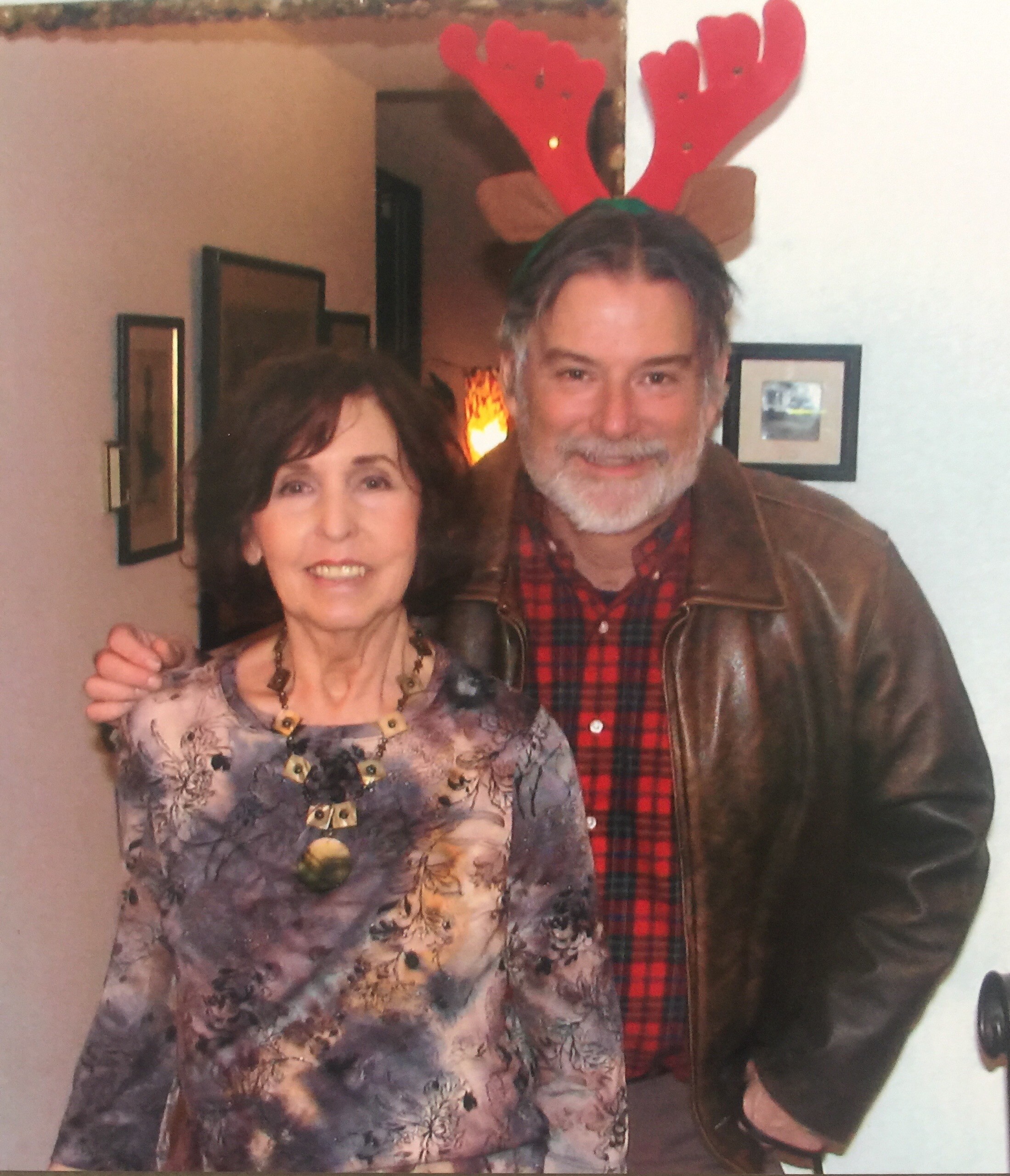 Smiling woman in floral blouse with necklace posing next to a man wearing red plaid shirt and reindeer antlers indoors.