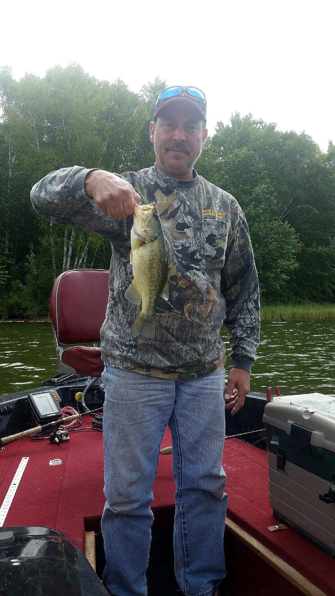 Man in camo shirt on a boat proudly holds up a largemouth bass he just caught on a lake.