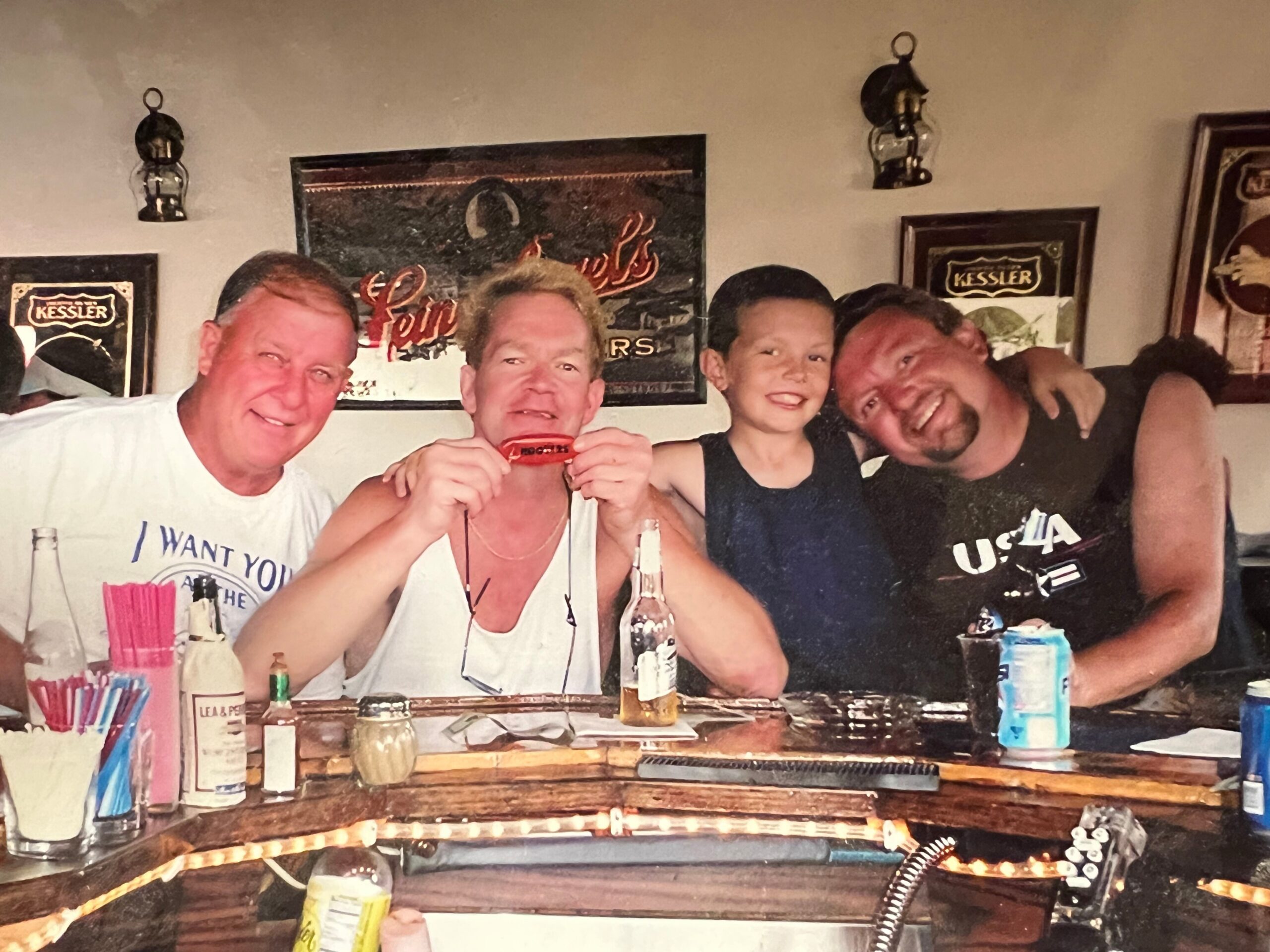 Group of friends at a bar posing for a photo with drinks on the counter and wall art in the background.