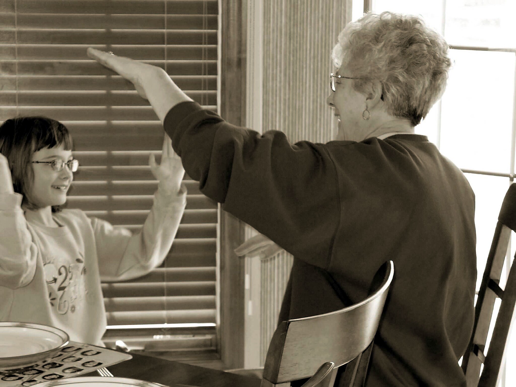 Older woman with short gray hair and glasses points at window blinds while a smiling child watches in a kitchen.